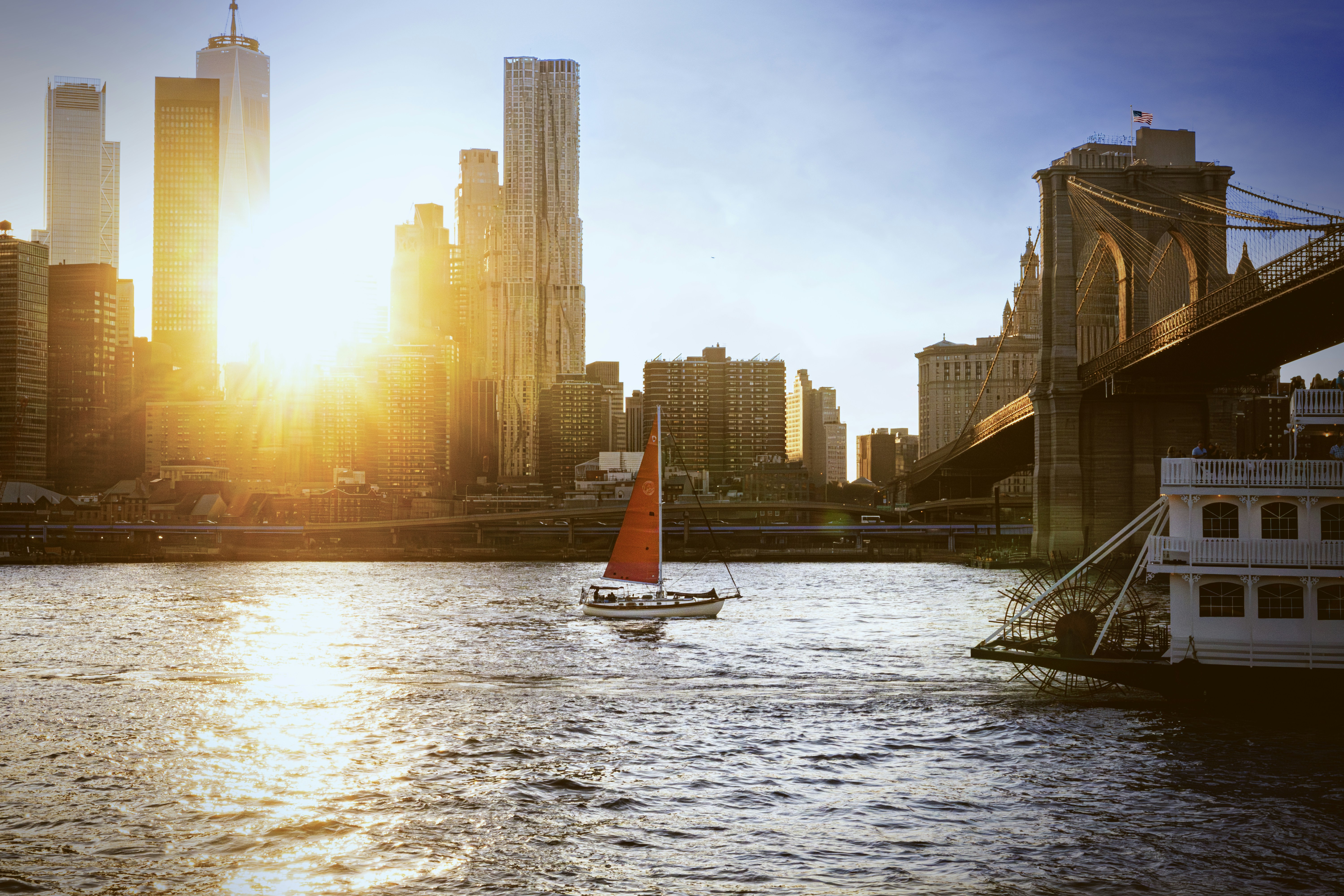 A sailboat with an orange sail near the Brooklyn Bridge in New York
