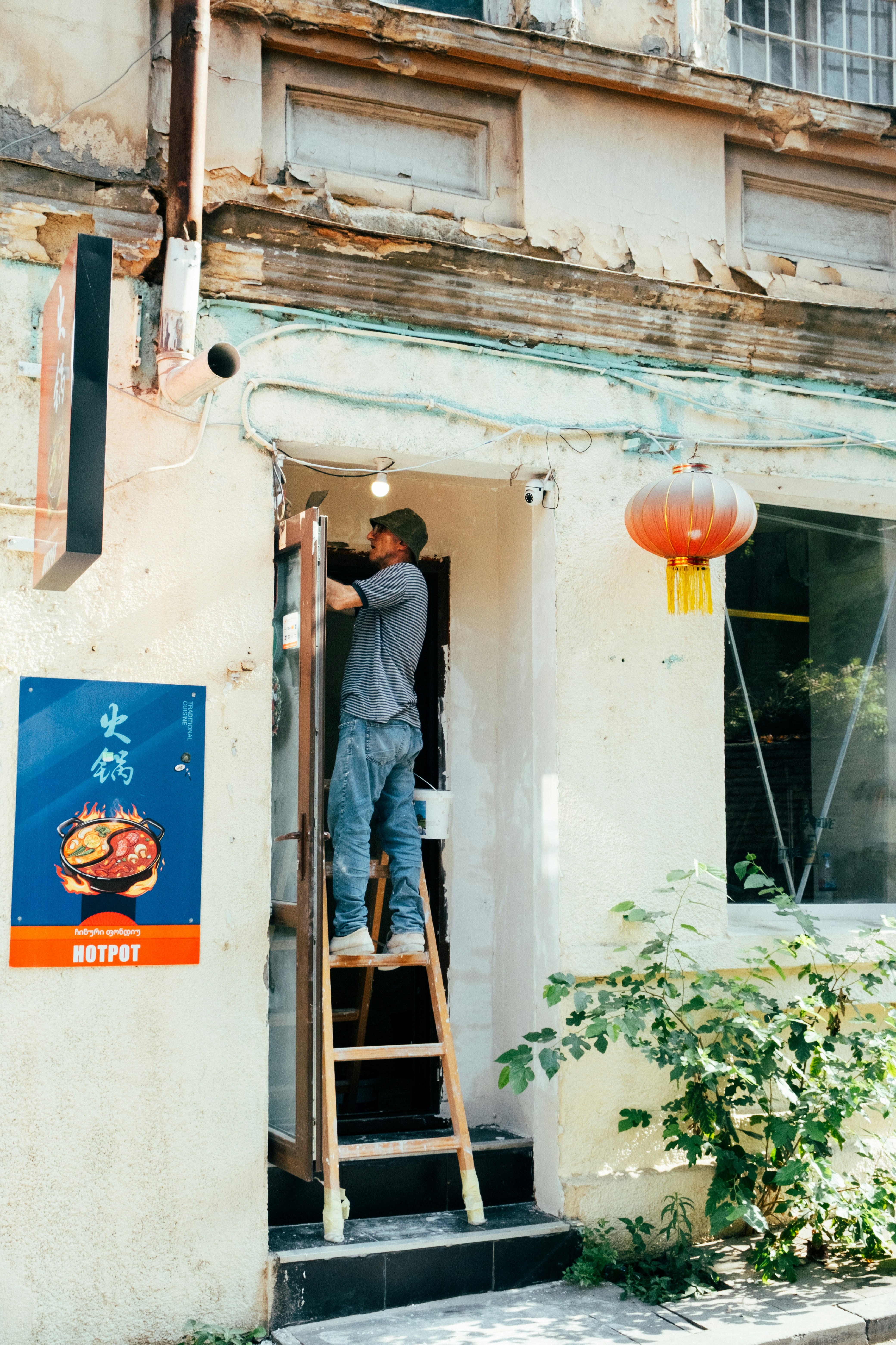 A person on a wooden ladder painting a doorway near a hotpot sign