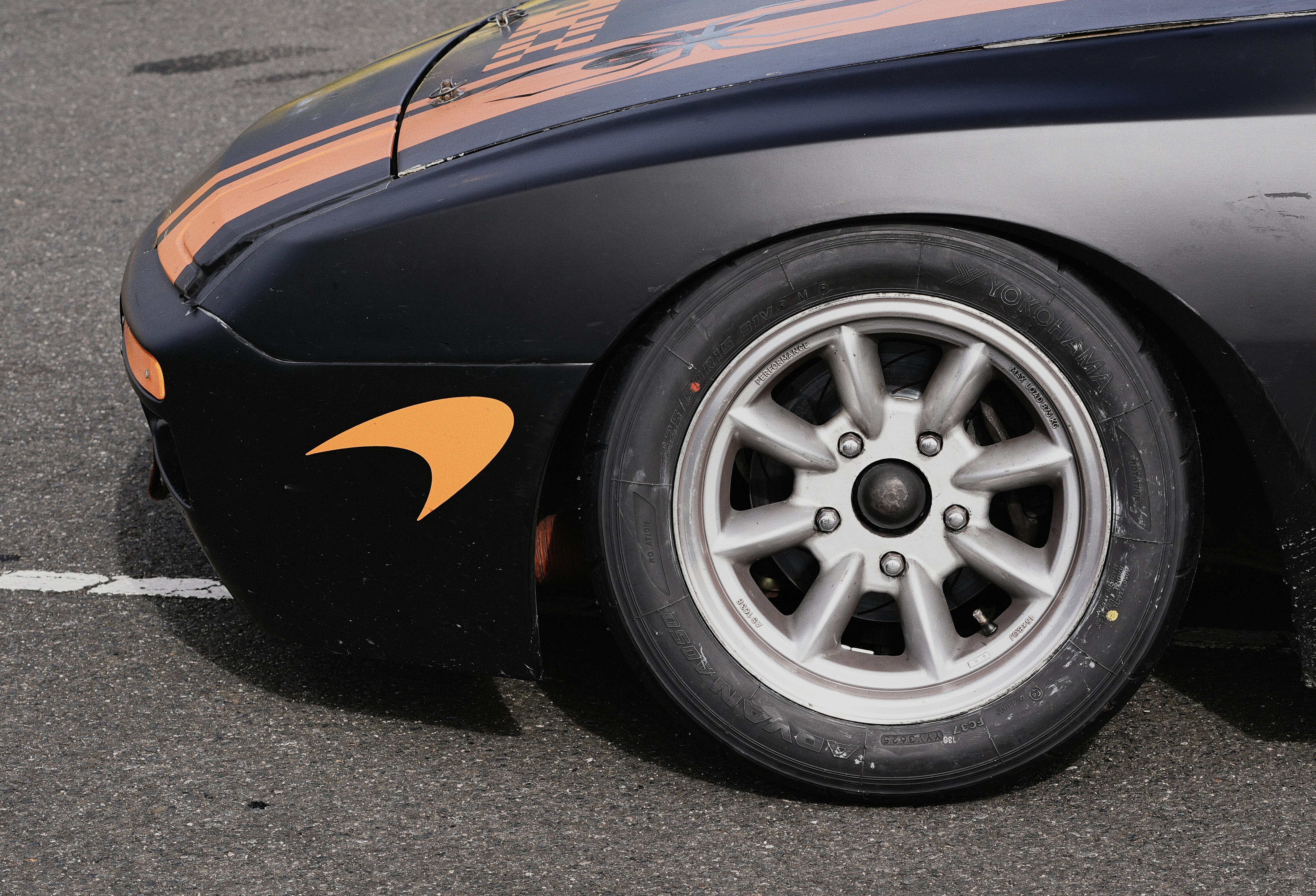 A black car with orange racing stripes and a silver wheel on asphalt