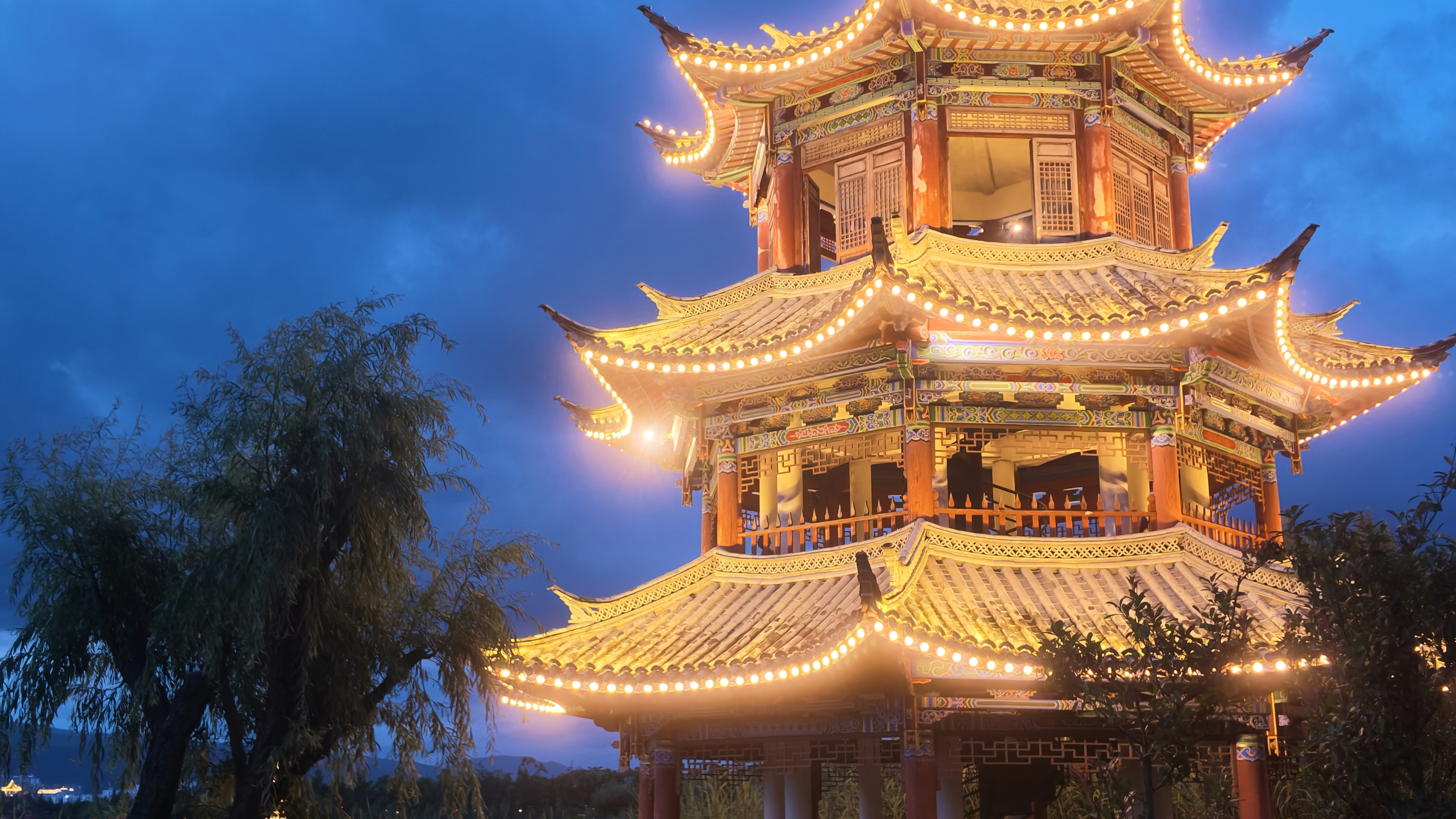 Tiered illuminated pagoda rises against a dusky blue sky with surrounding trees and soft foreground silhouettes.