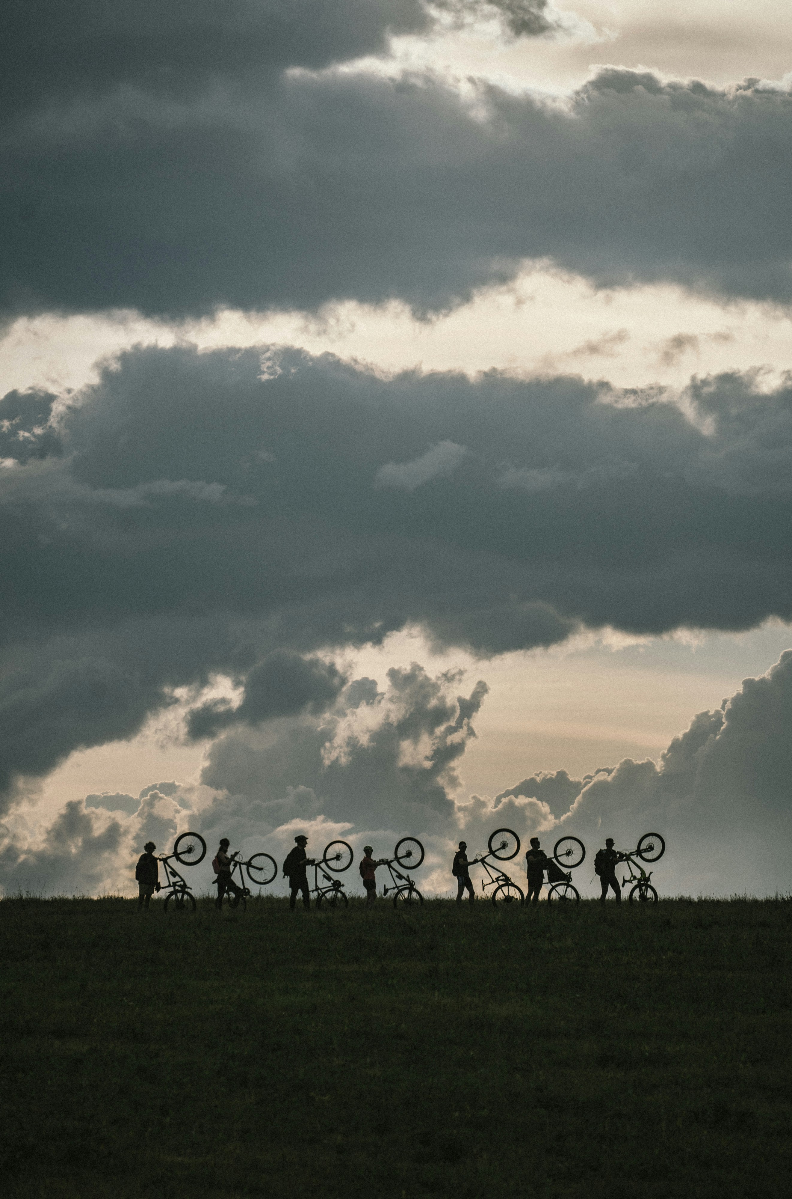 A group of people in silhouette carrying bicycles across a grassy hill under clouds