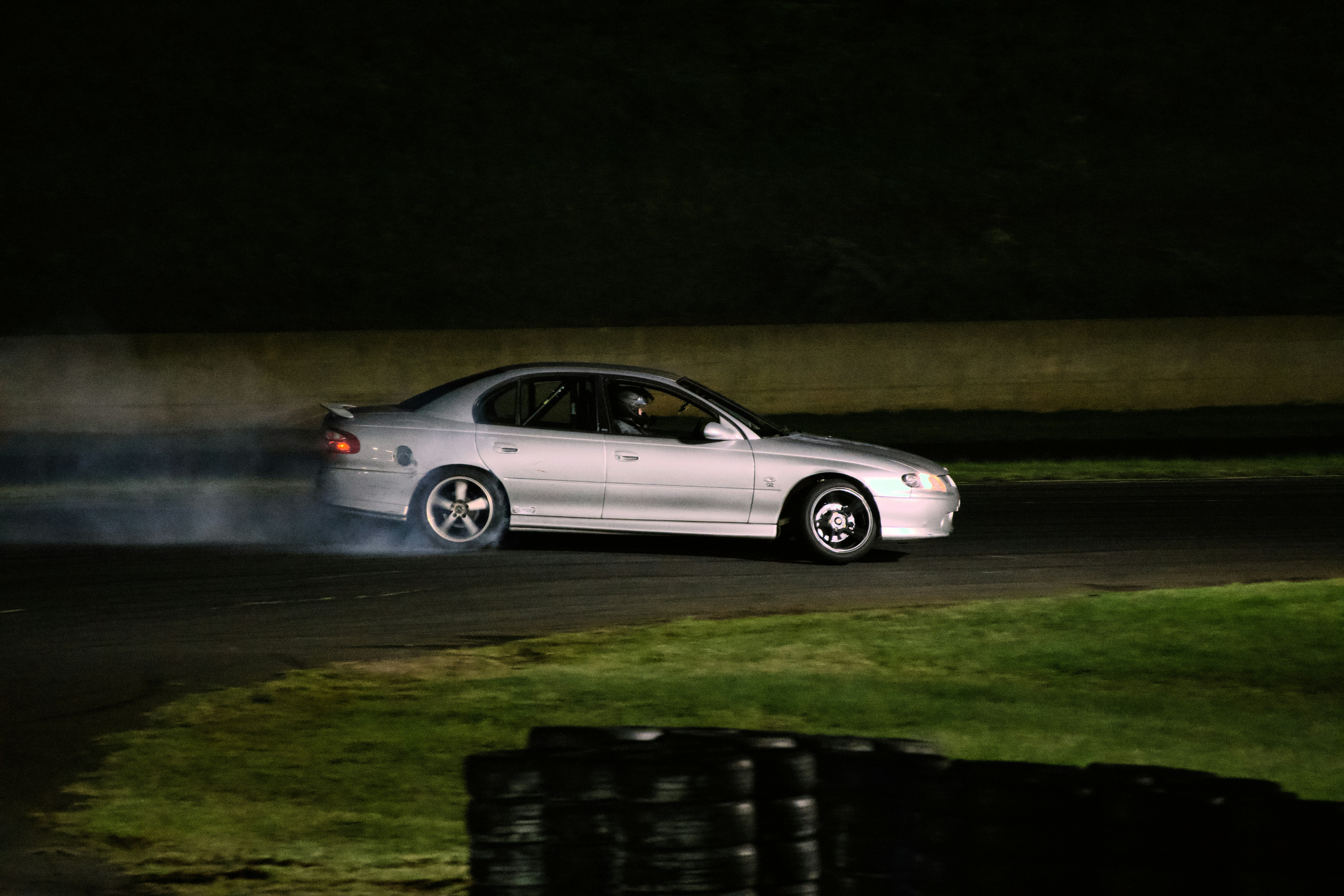 A silver sedan drifting on a race track at night with tire smoke