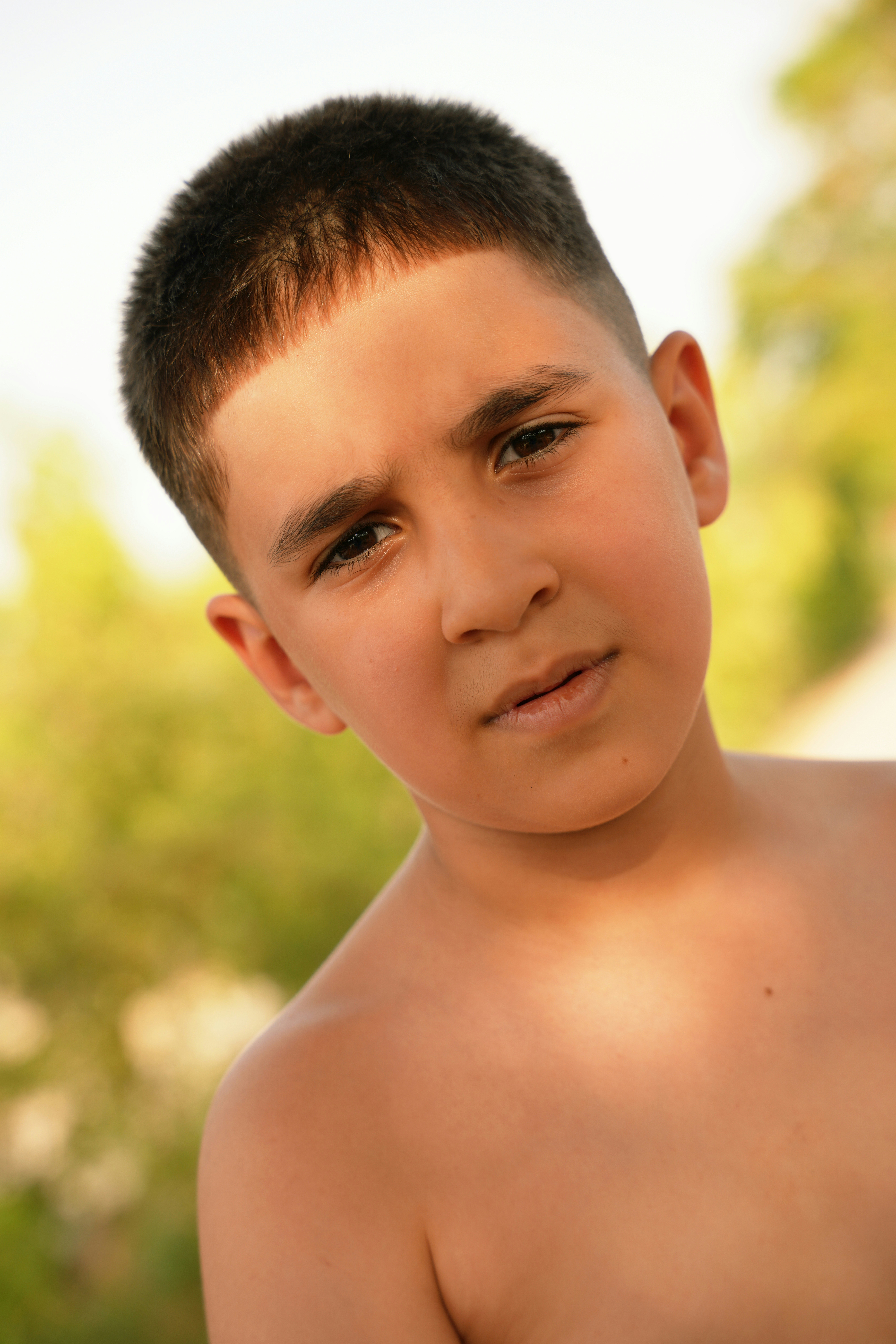 A shirtless boy with a buzz cut squints softly in warm outdoor light