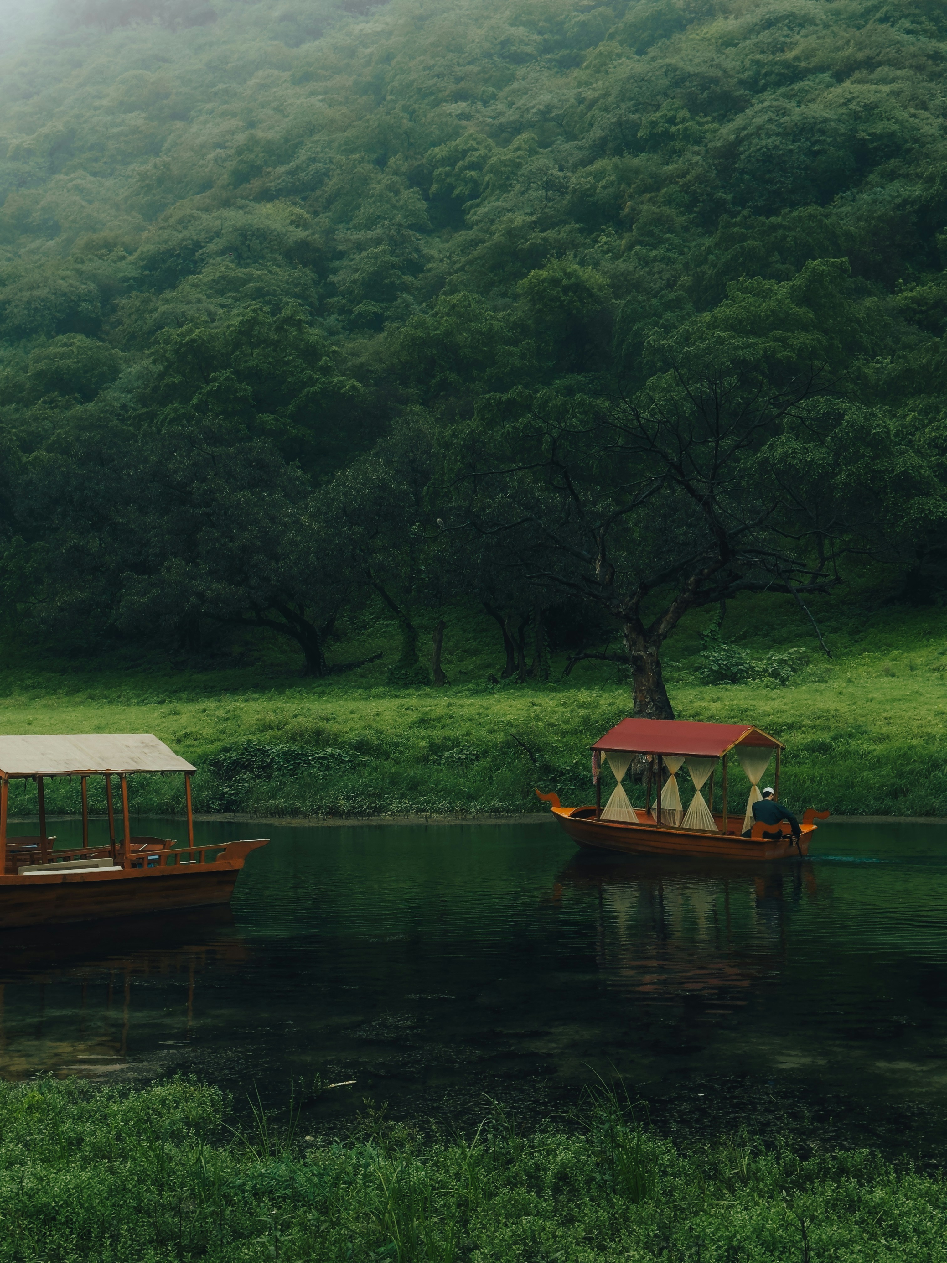 Two wooden boats on a calm river beside a lush green forested hillside