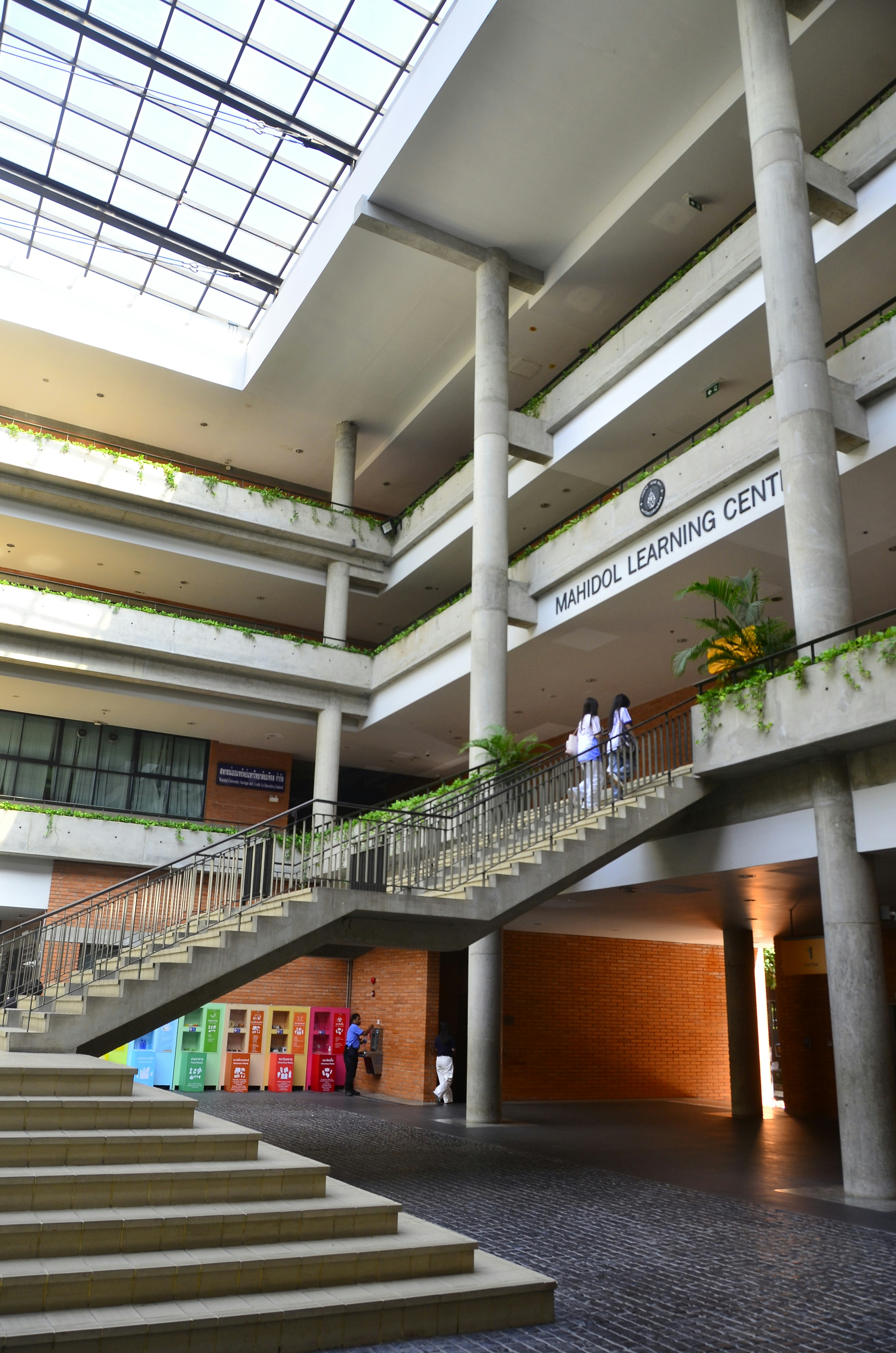 People walking up a concrete staircase inside the Mahidol Learning Center with a glass roof