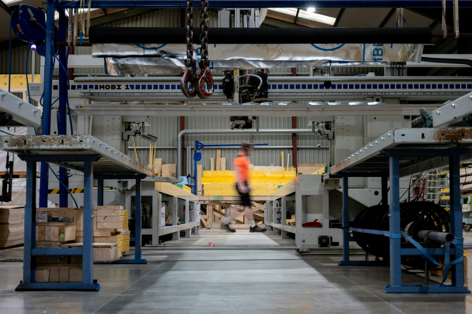 A long fabrication bay with overhead crane rails and work benches, a worker in an orange shirt at the far end.