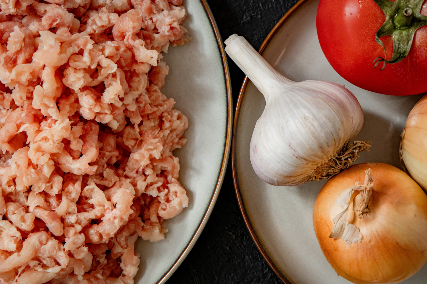 Raw ground meat on a plate beside garlic, onions, and a tomato