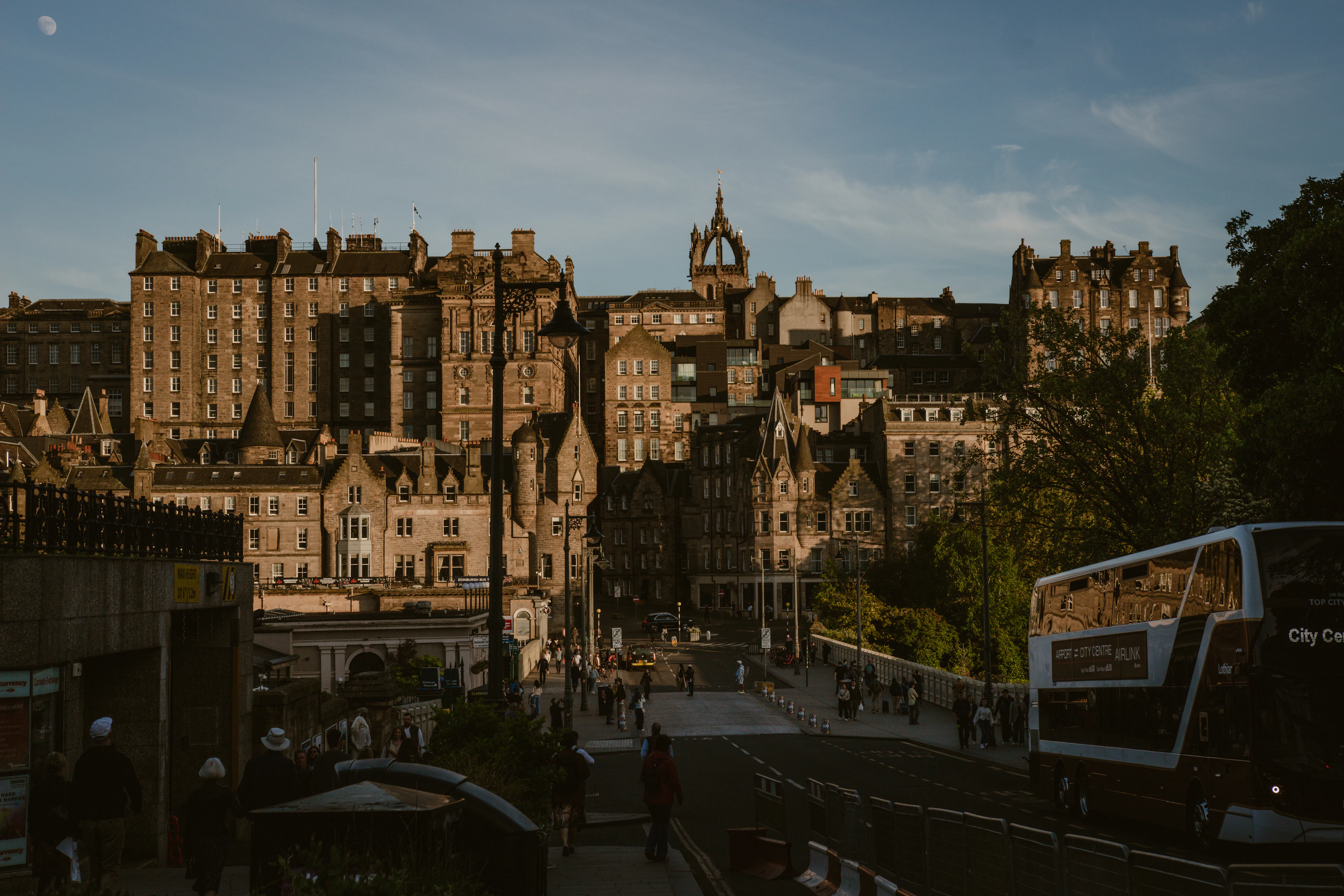 The tenements, crow-stepped gables and spires of Edinburgh's Old Town stacked up in warm evening light above Princes Street, with an open-top double-decker bus waiting at the kerb in the foreground.