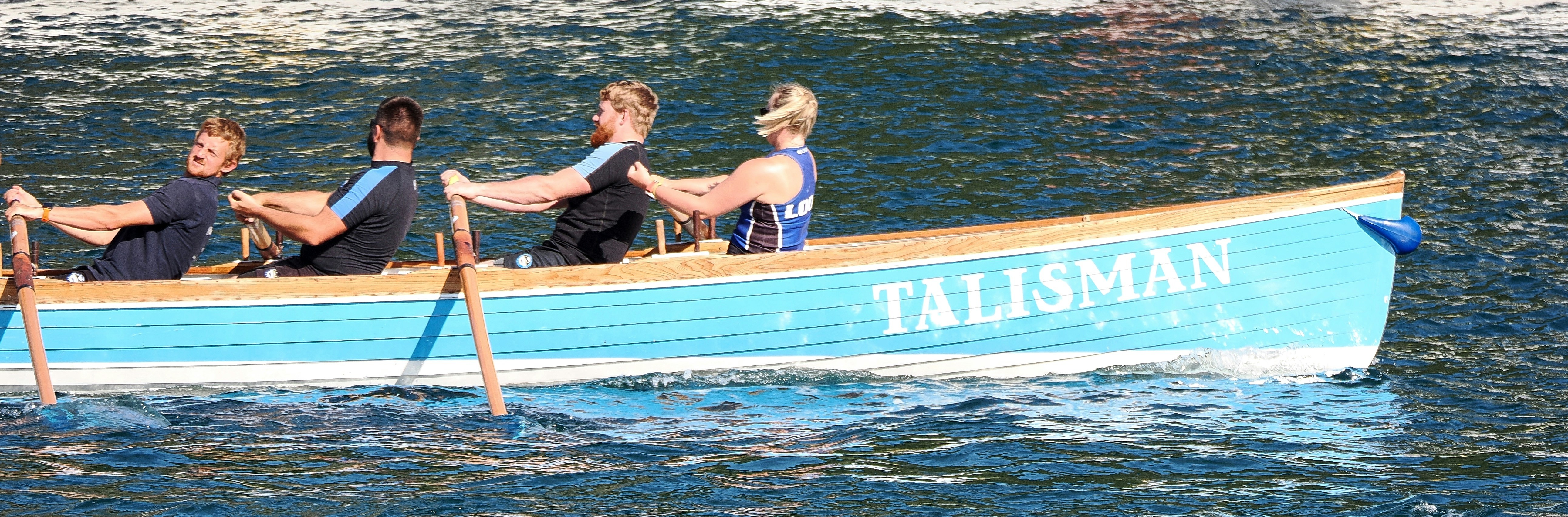 Four people rowing a light blue boat named Talisman on dark water