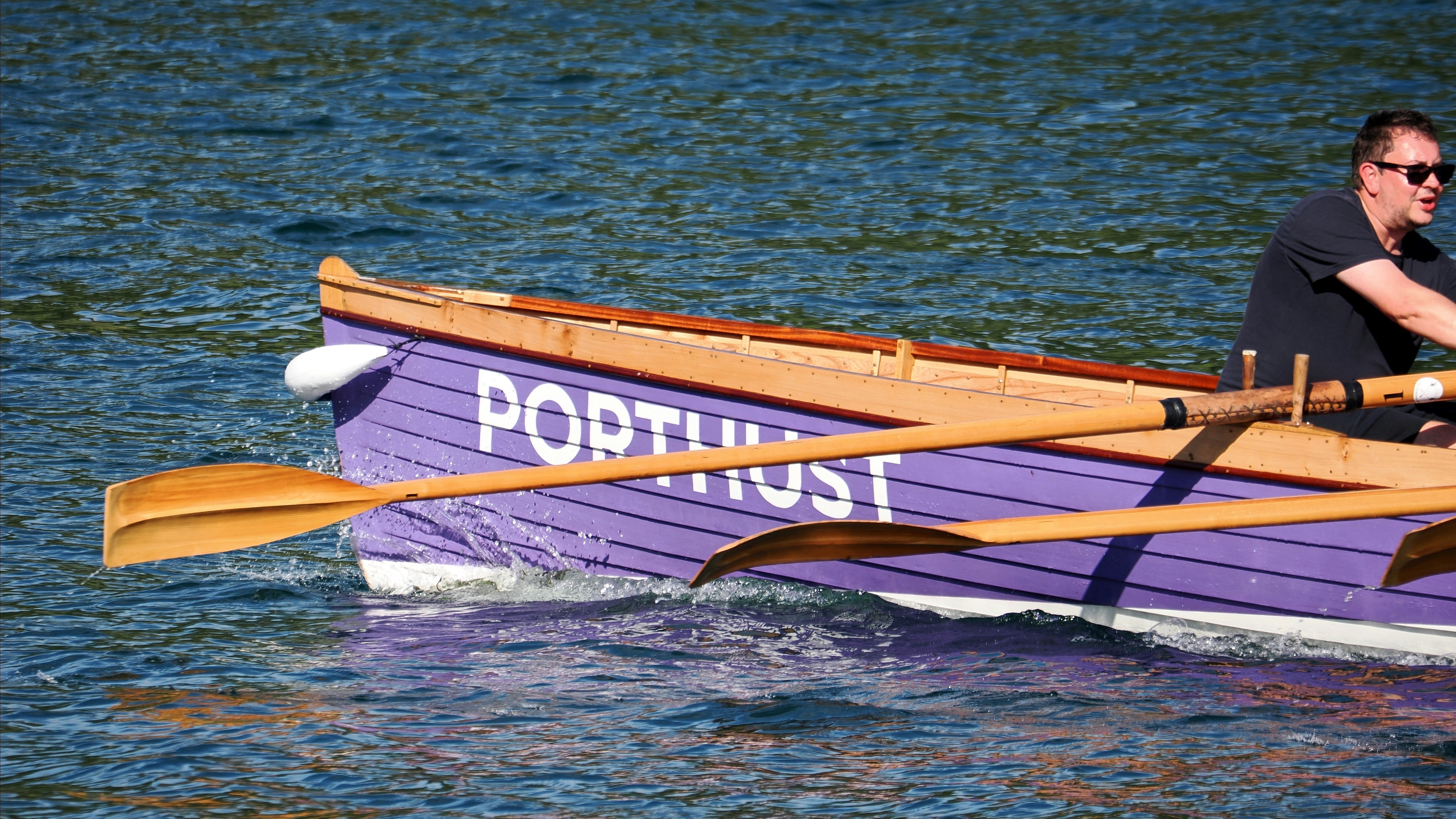 A man rowing a purple wooden boat labeled PORTHUST on blue water