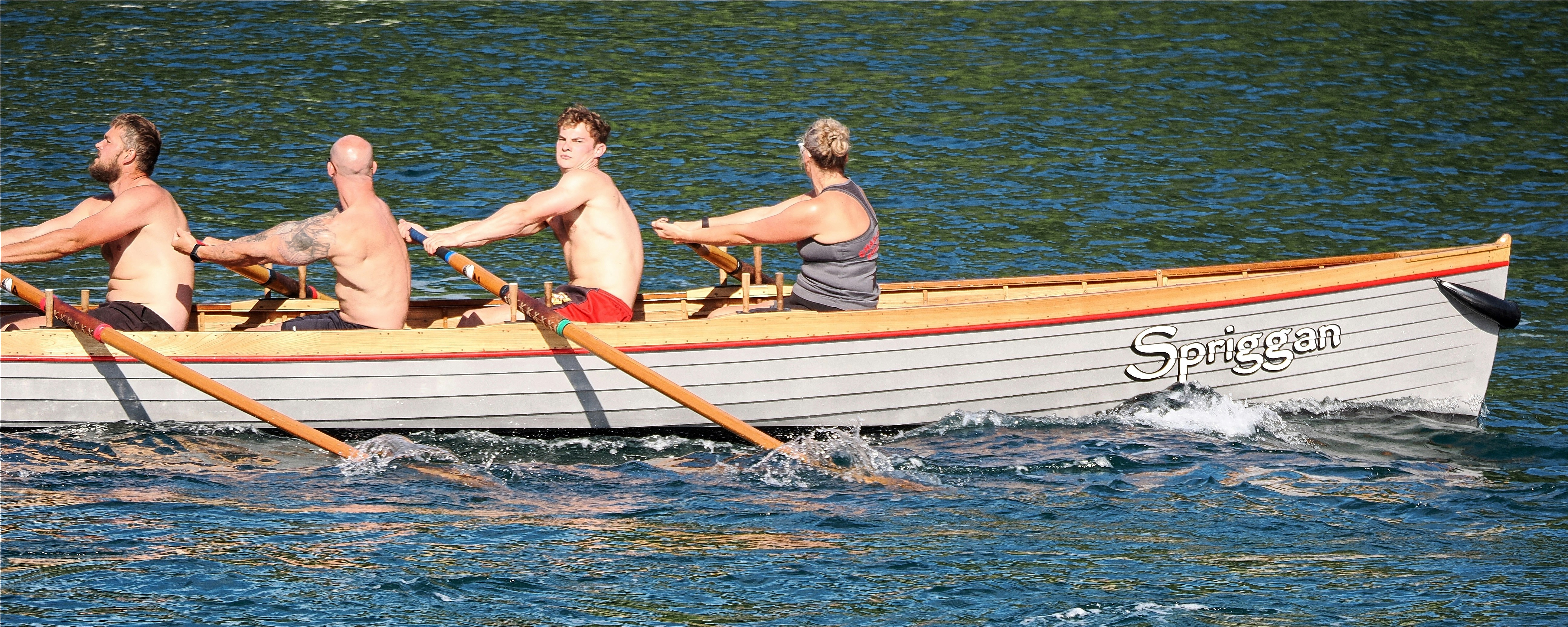 Four people rowing a wooden boat named Spriggan on blue water
