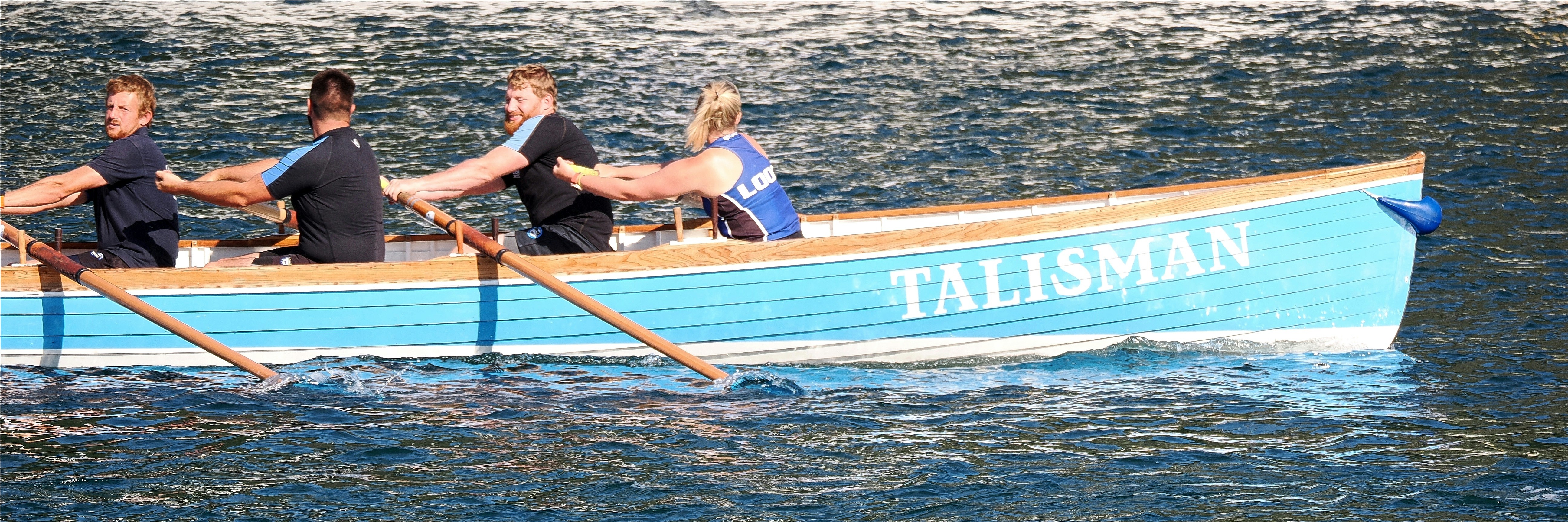 Four people rowing a light blue boat named Talisman on dark water