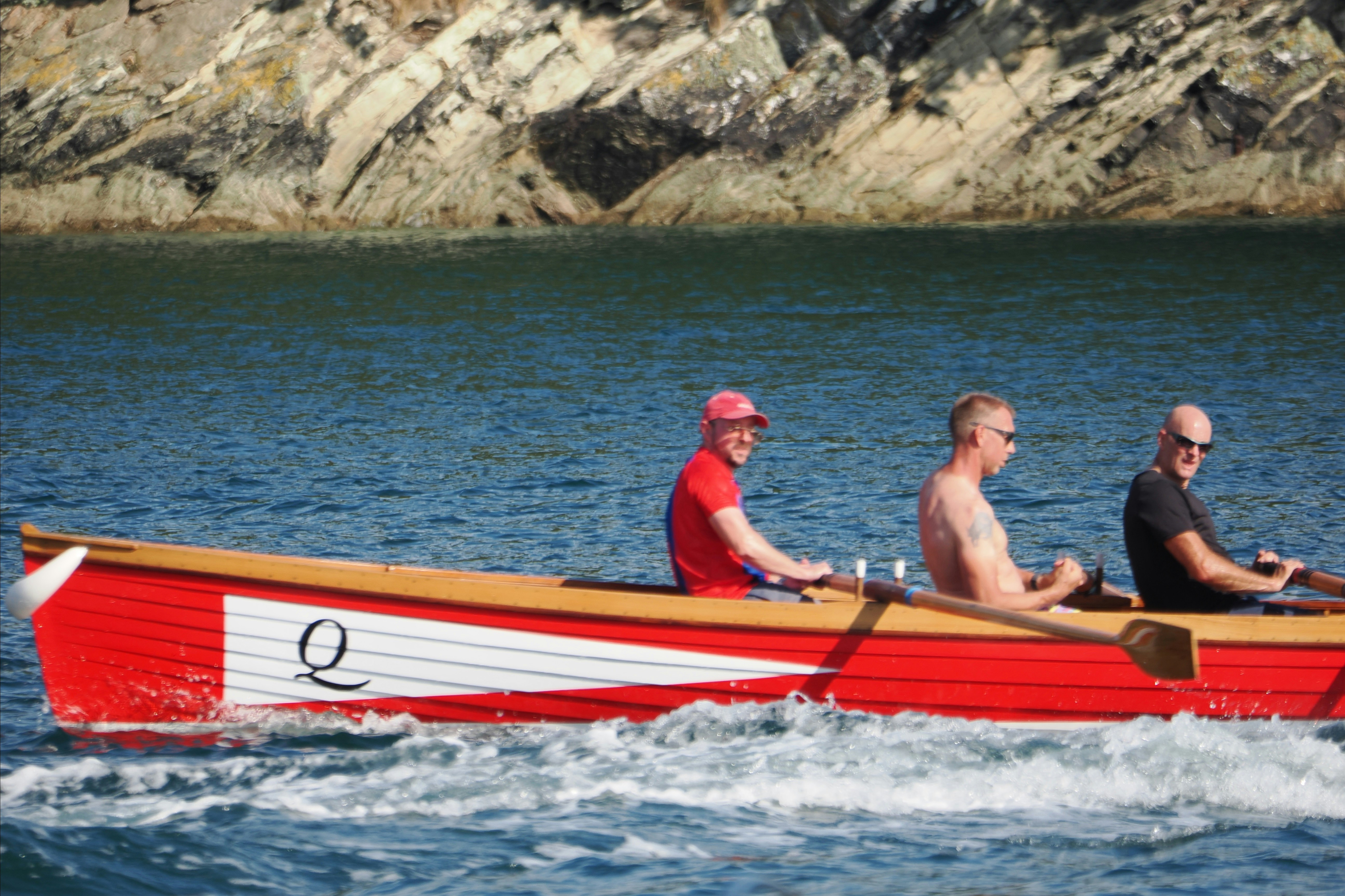 Three men rowing a red wooden boat with a white stripe on blue water