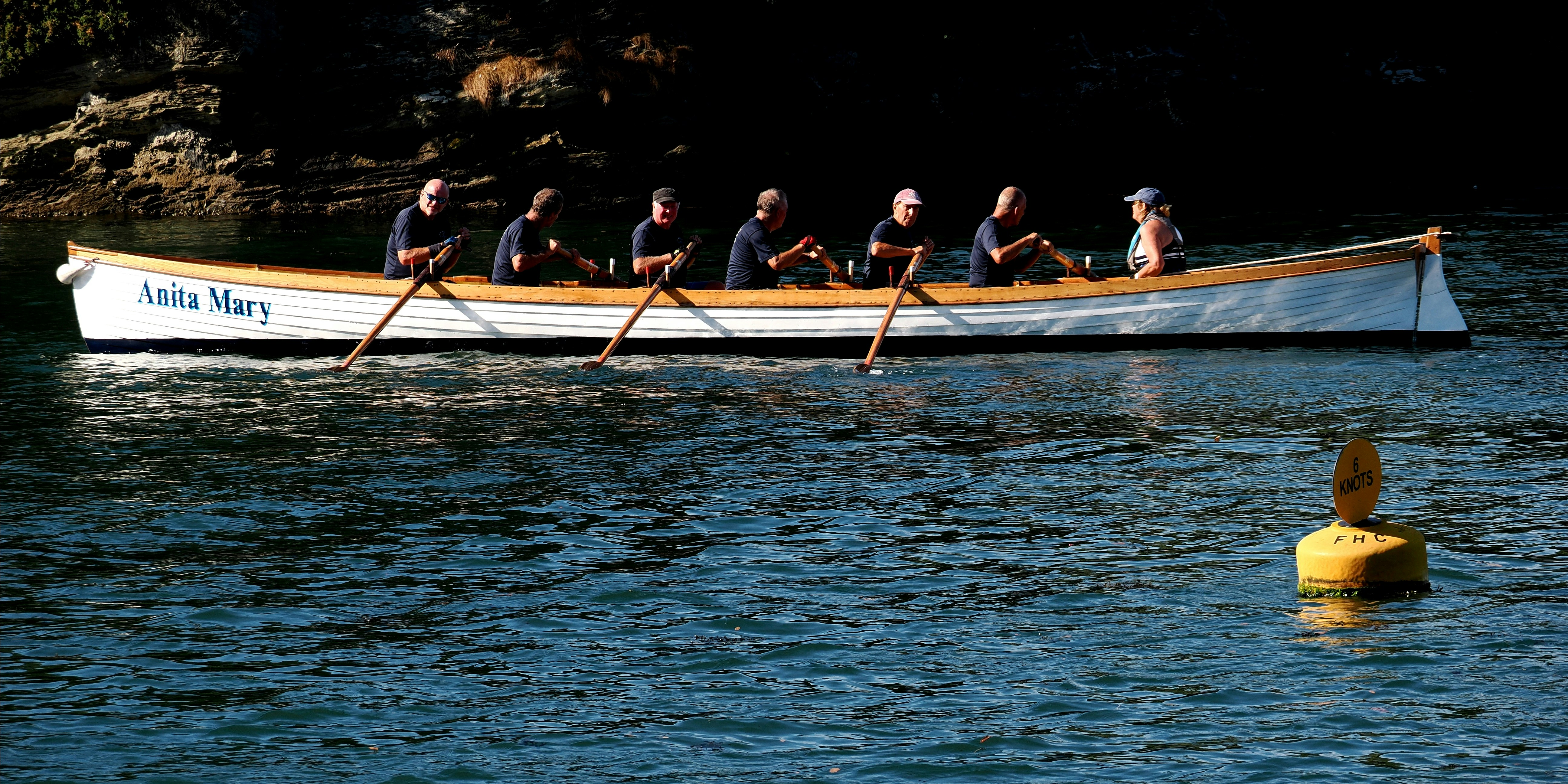 A group of people rowing a long white boat named Anita Mary on water