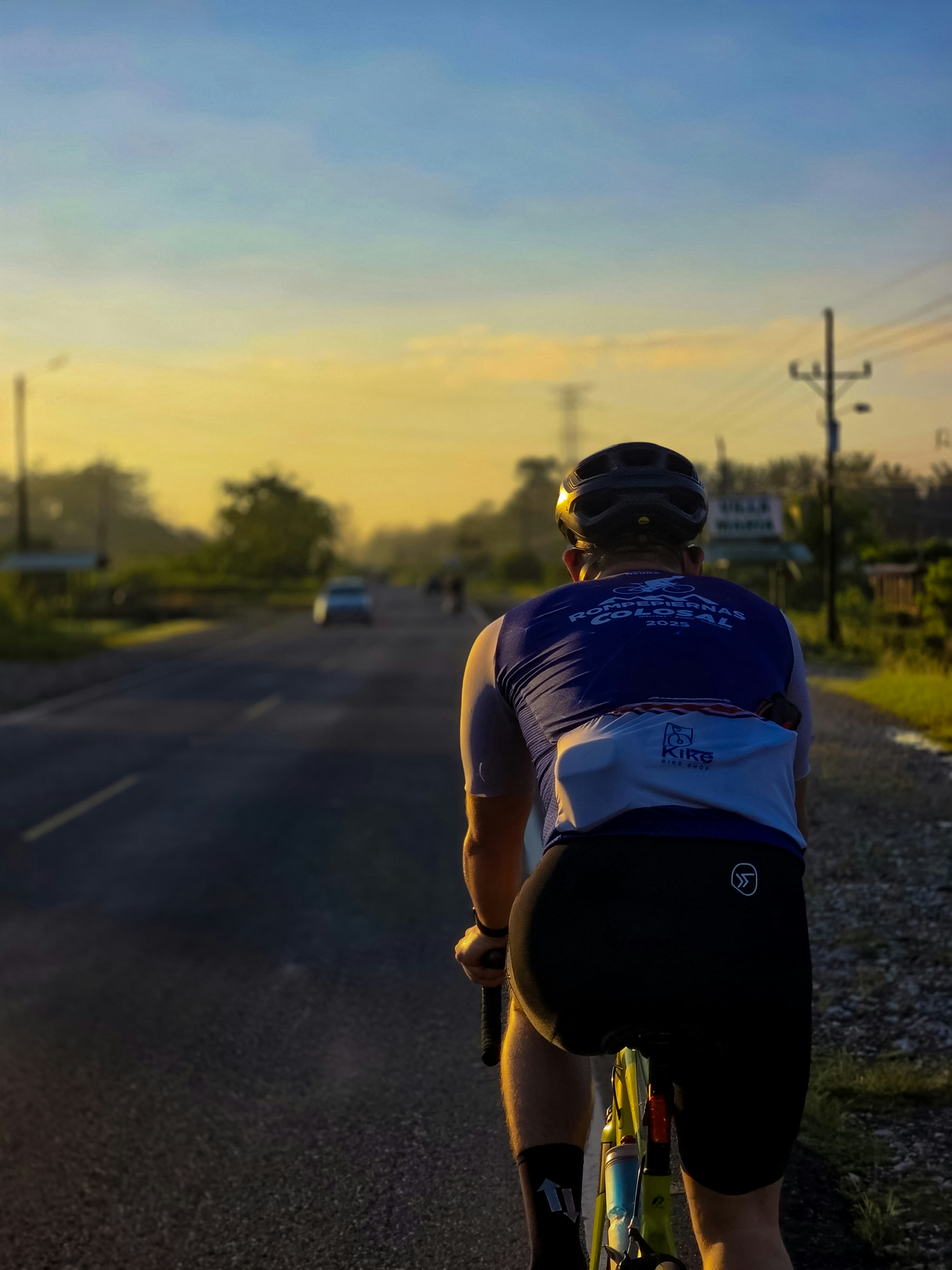 A cyclist in a blue jersey and helmet riding on a road at golden hour