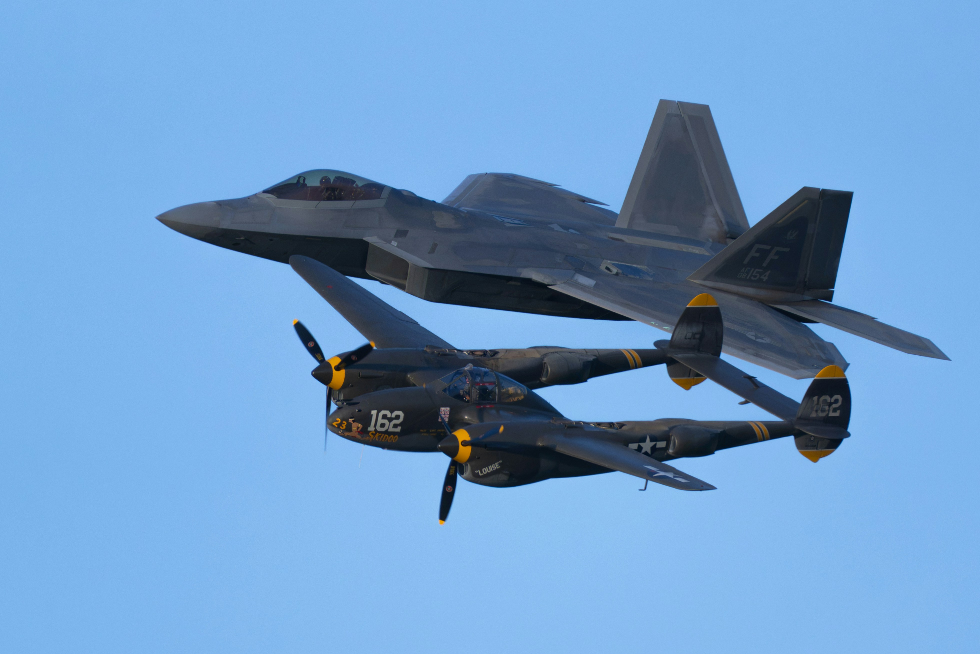 A modern stealth fighter flying in formation with a vintage twin-engine propeller plane