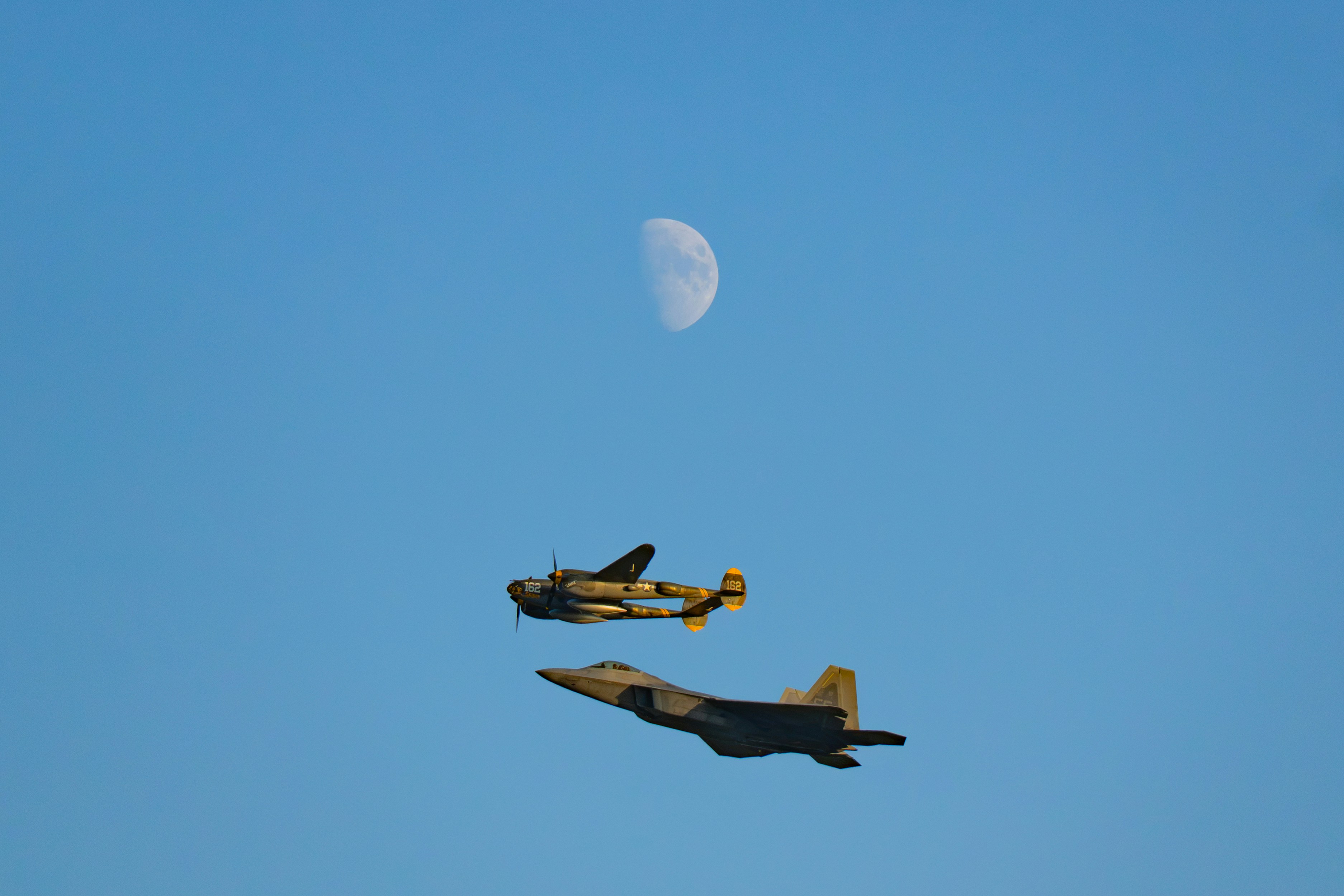 A vintage propeller plane and a modern fighter jet flying beneath a half moon