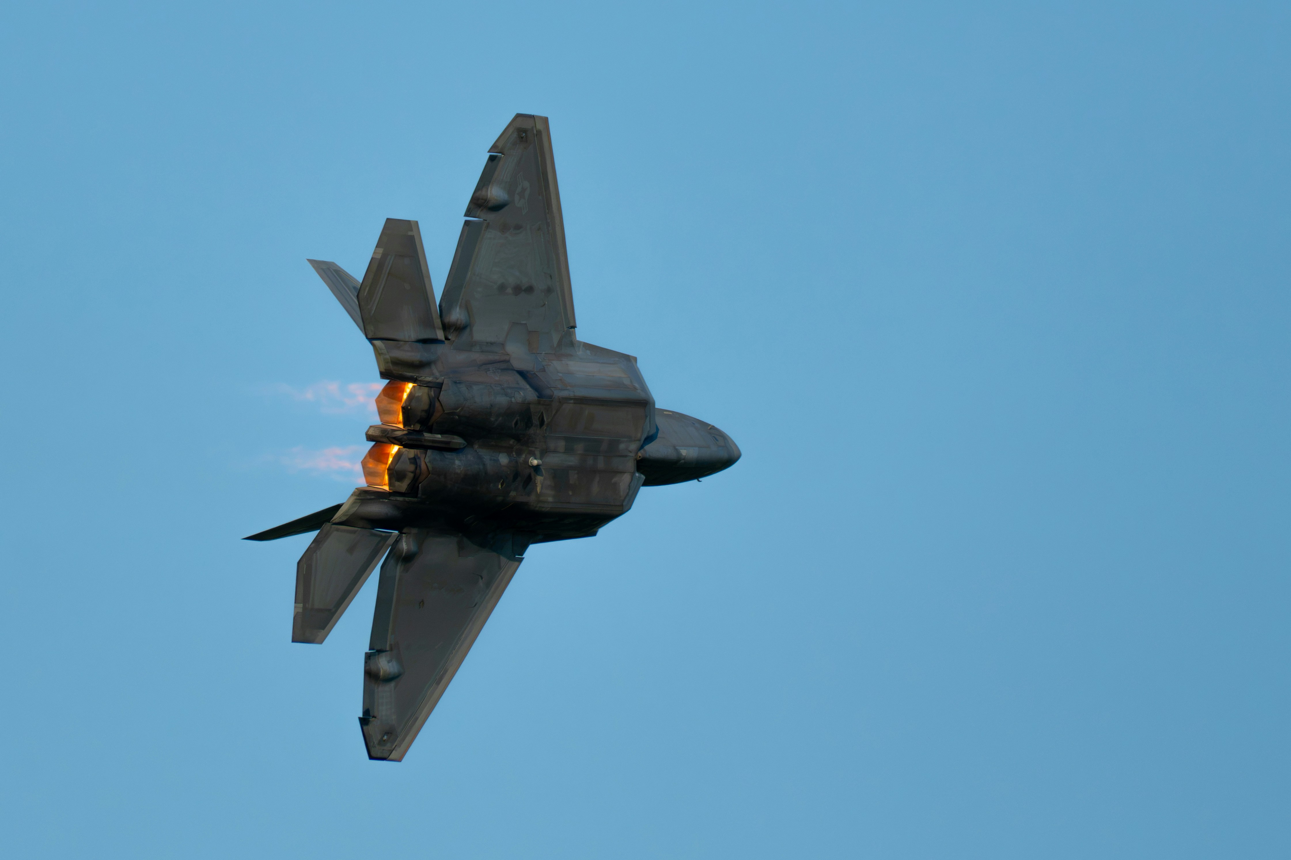 A grey fighter jet climbing steeply against a clear blue sky with glowing exhaust