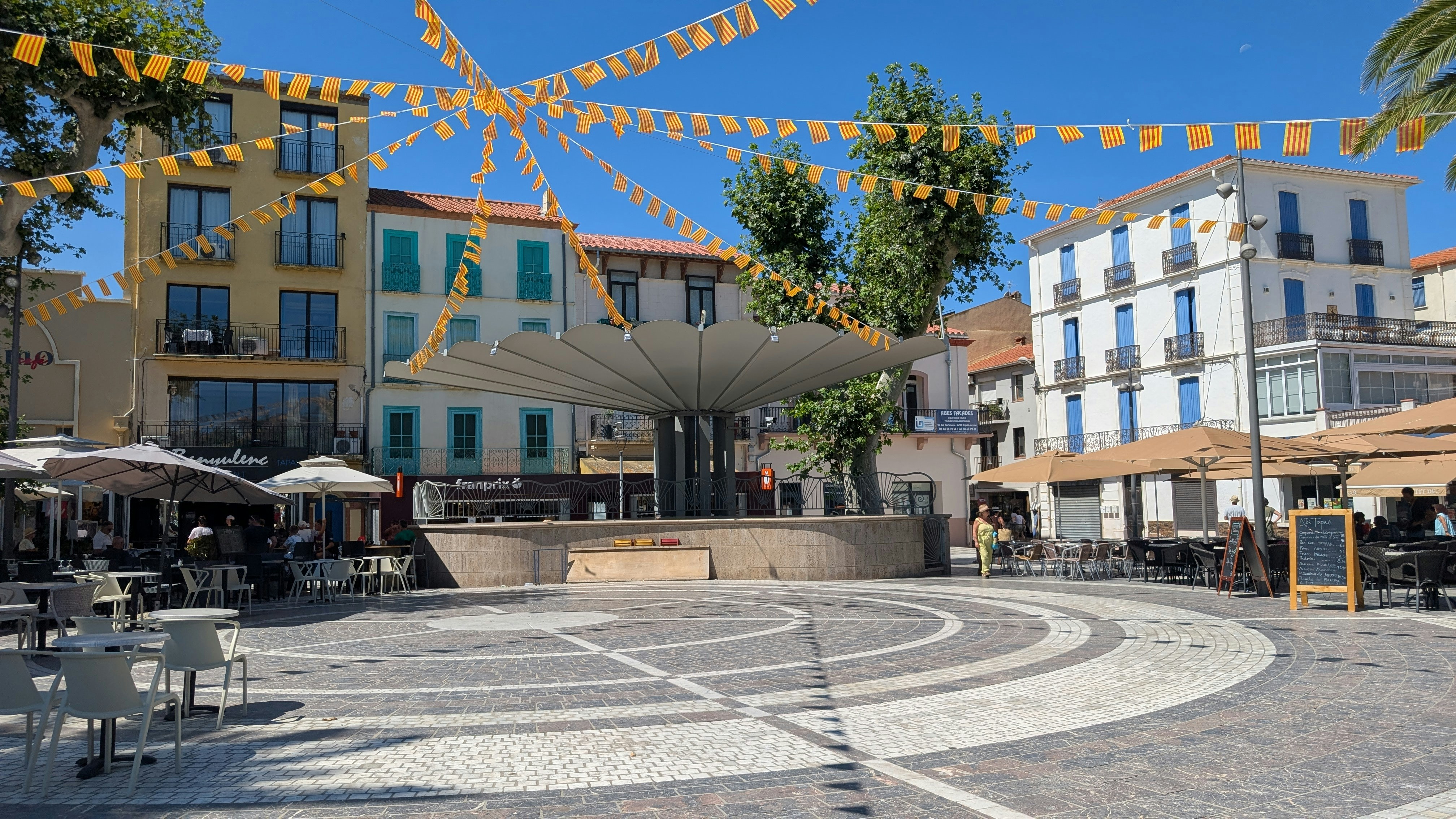 A town square with a large umbrella structure and yellow bunting under a blue sky