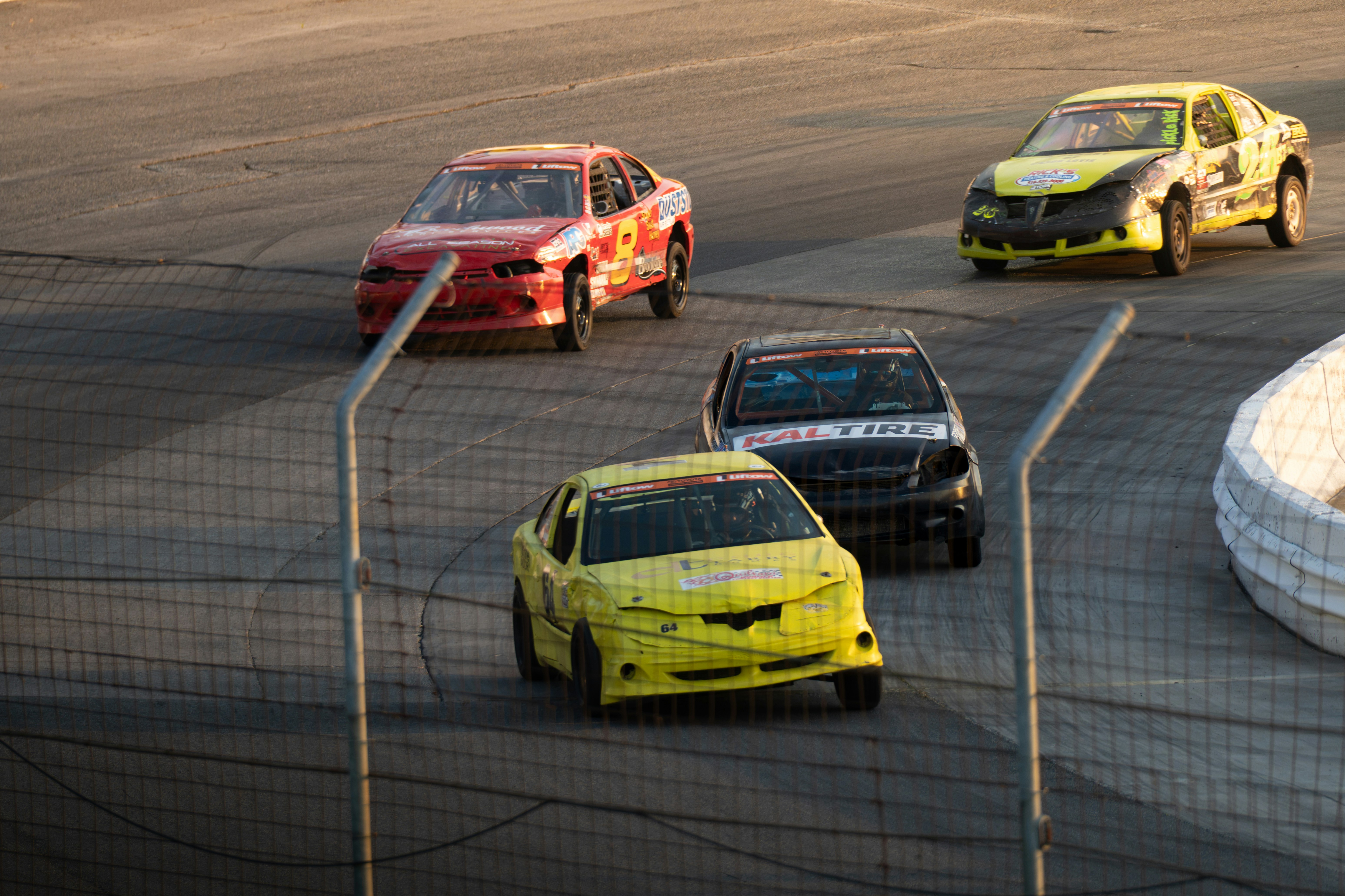 Four stock cars racing on an asphalt track behind a chain link fence