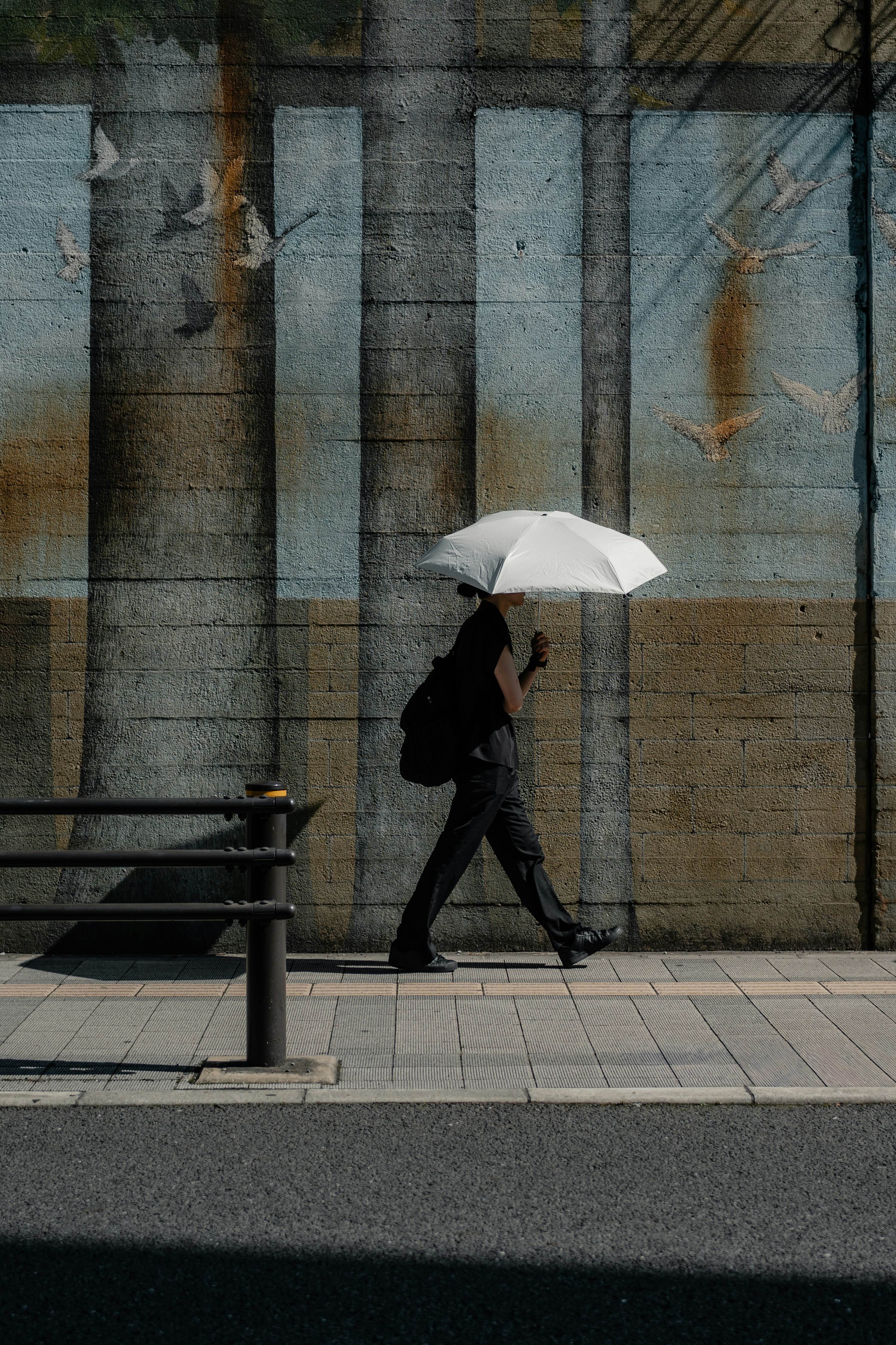 A person in black clothing walking with a white umbrella past a mural