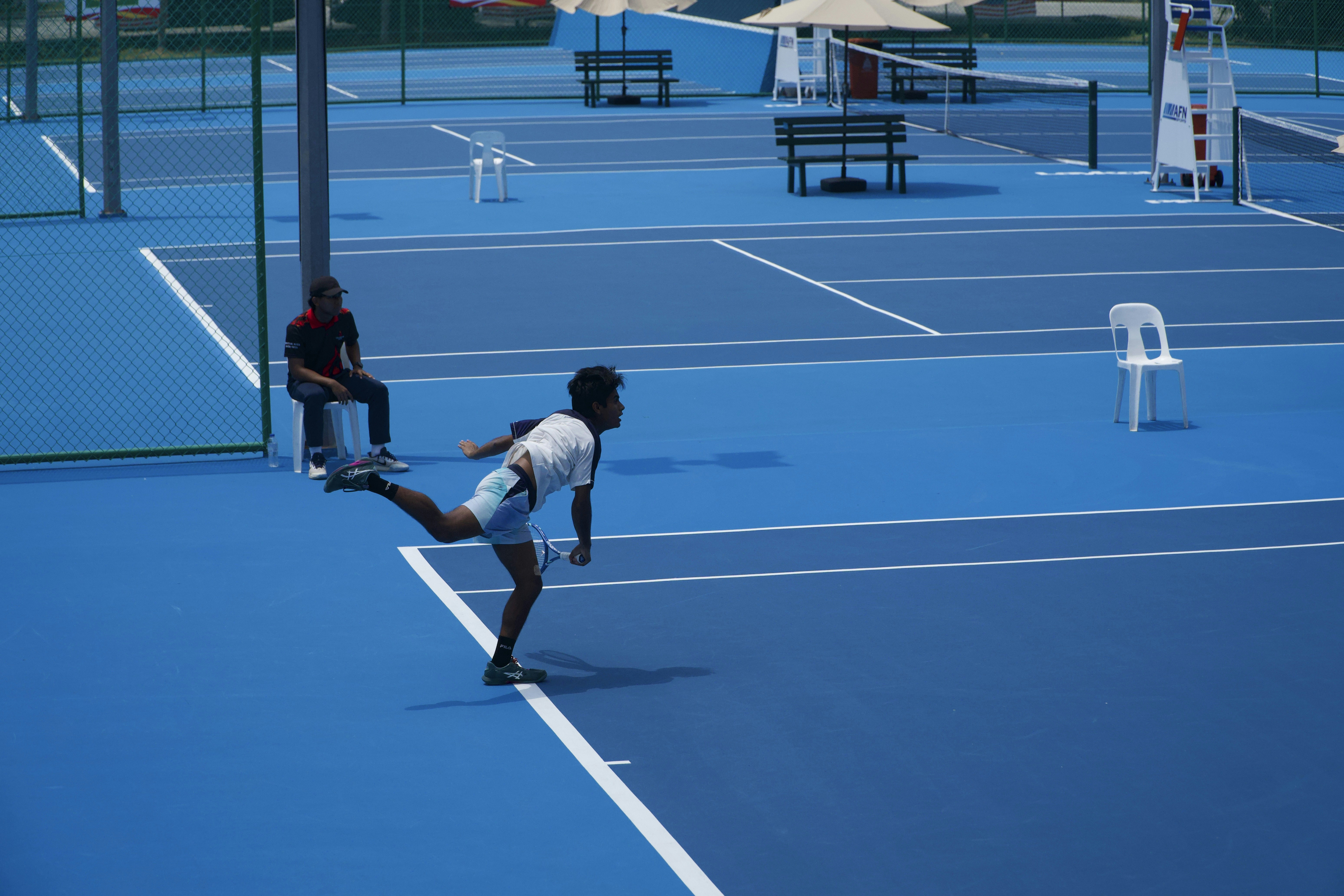 A tennis player serving on a bright blue court with a seated official
