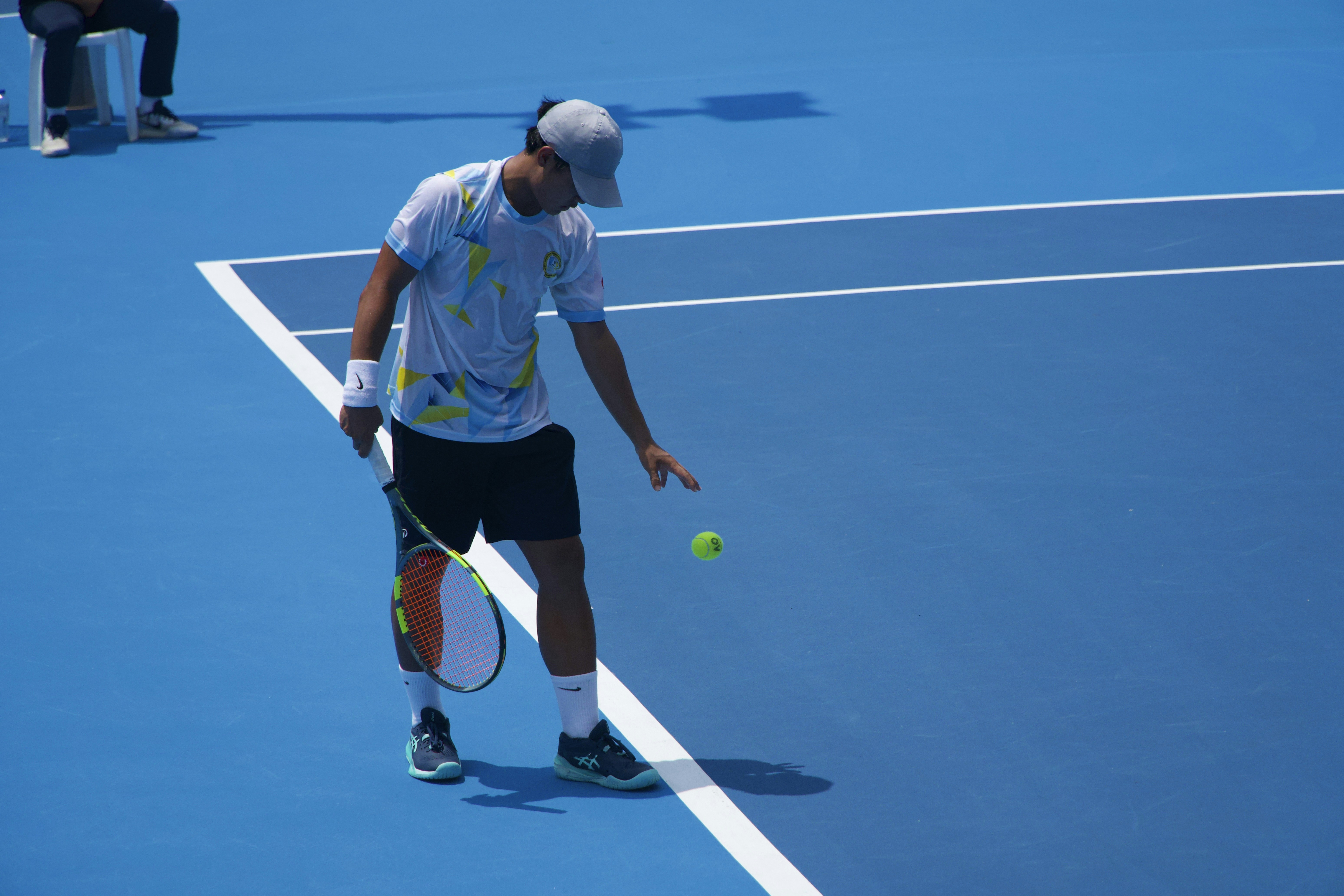 A tennis player bouncing a ball on a bright blue hard court
