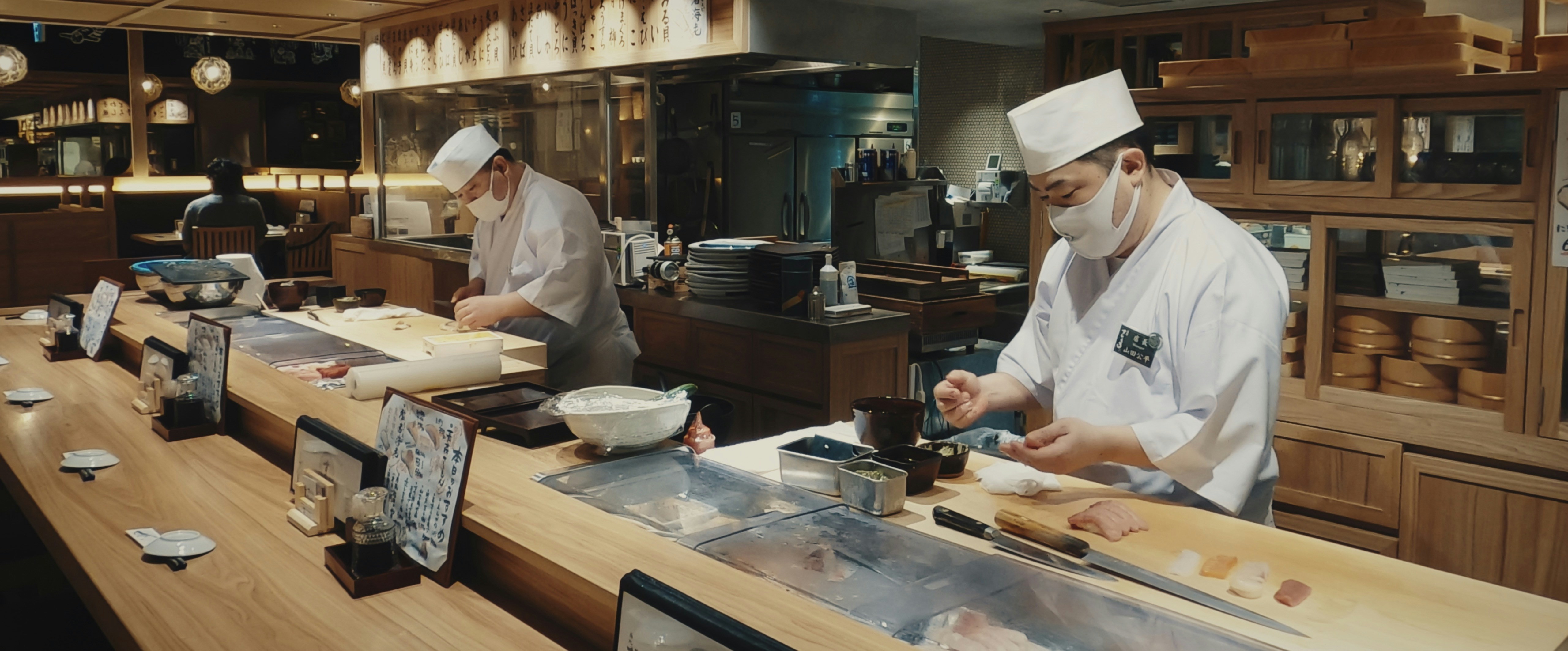 Two chefs in white uniforms and masks preparing sushi behind a wooden counter