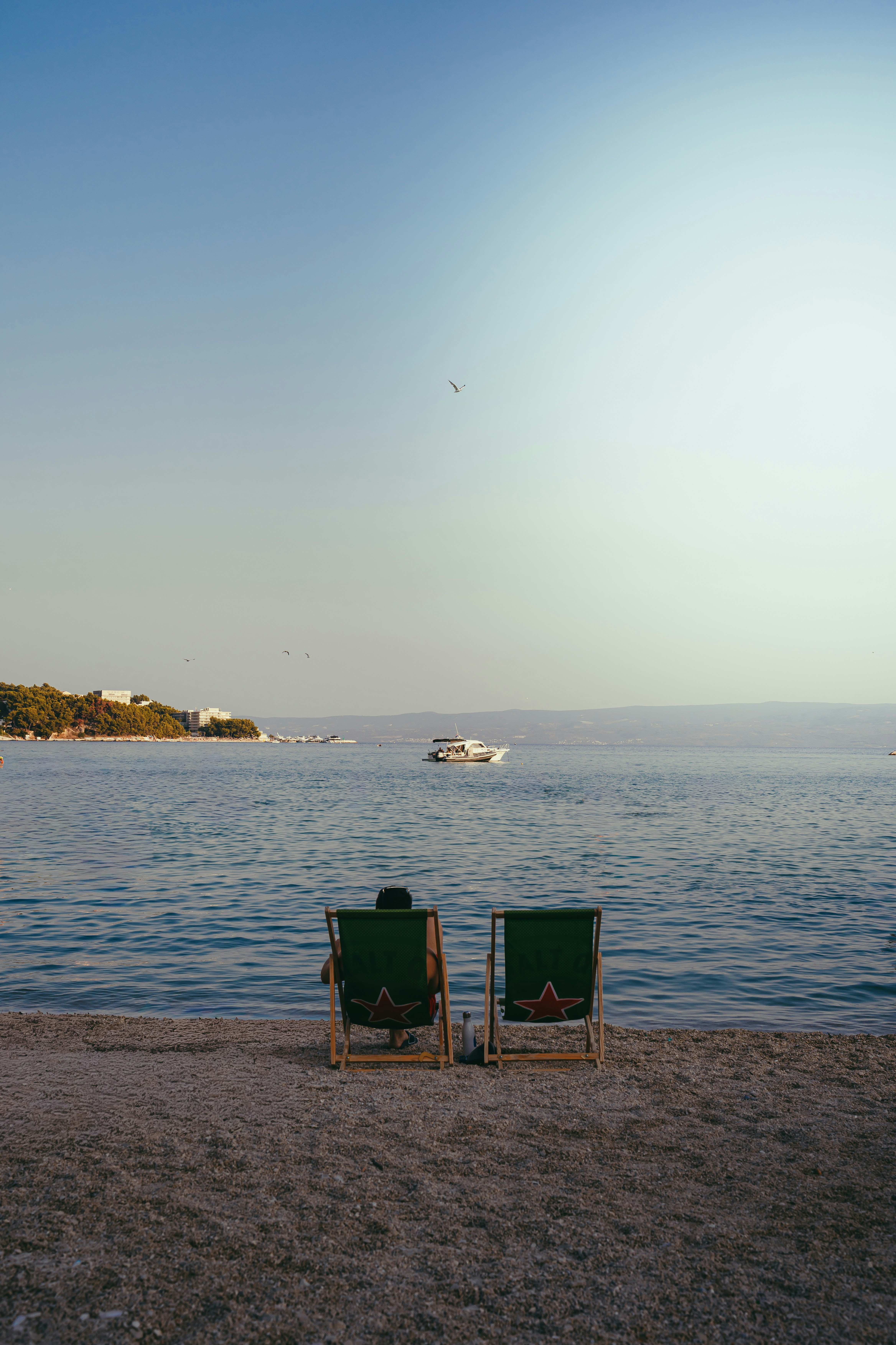 A person in a green deck chair on a pebble beach facing the sea