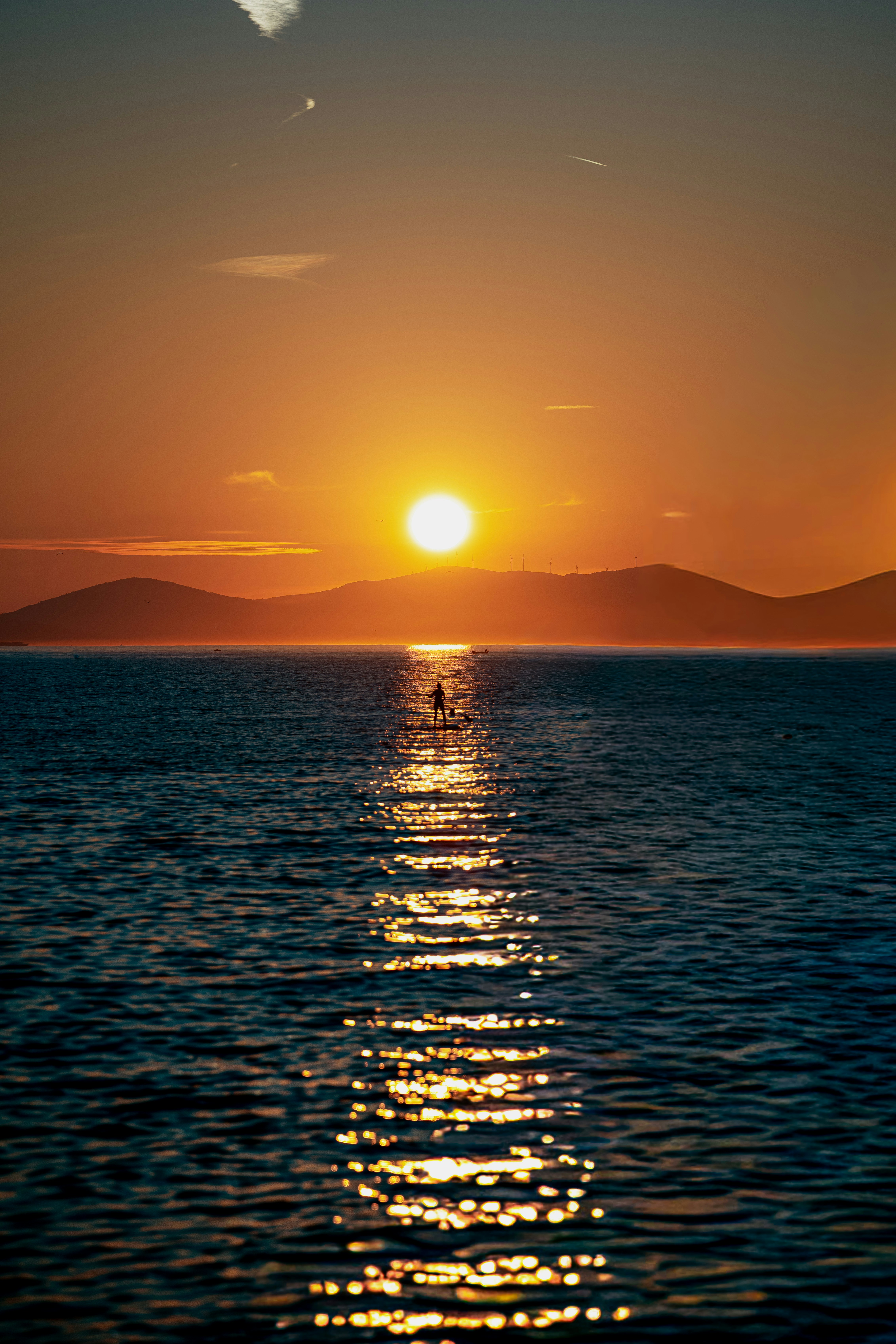 A person paddleboarding on dark water under a low golden sun and hills - 相关推荐