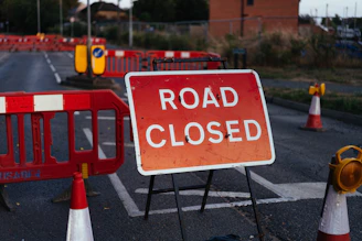A red road closed sign on a metal stand with orange barriers and traffic cones