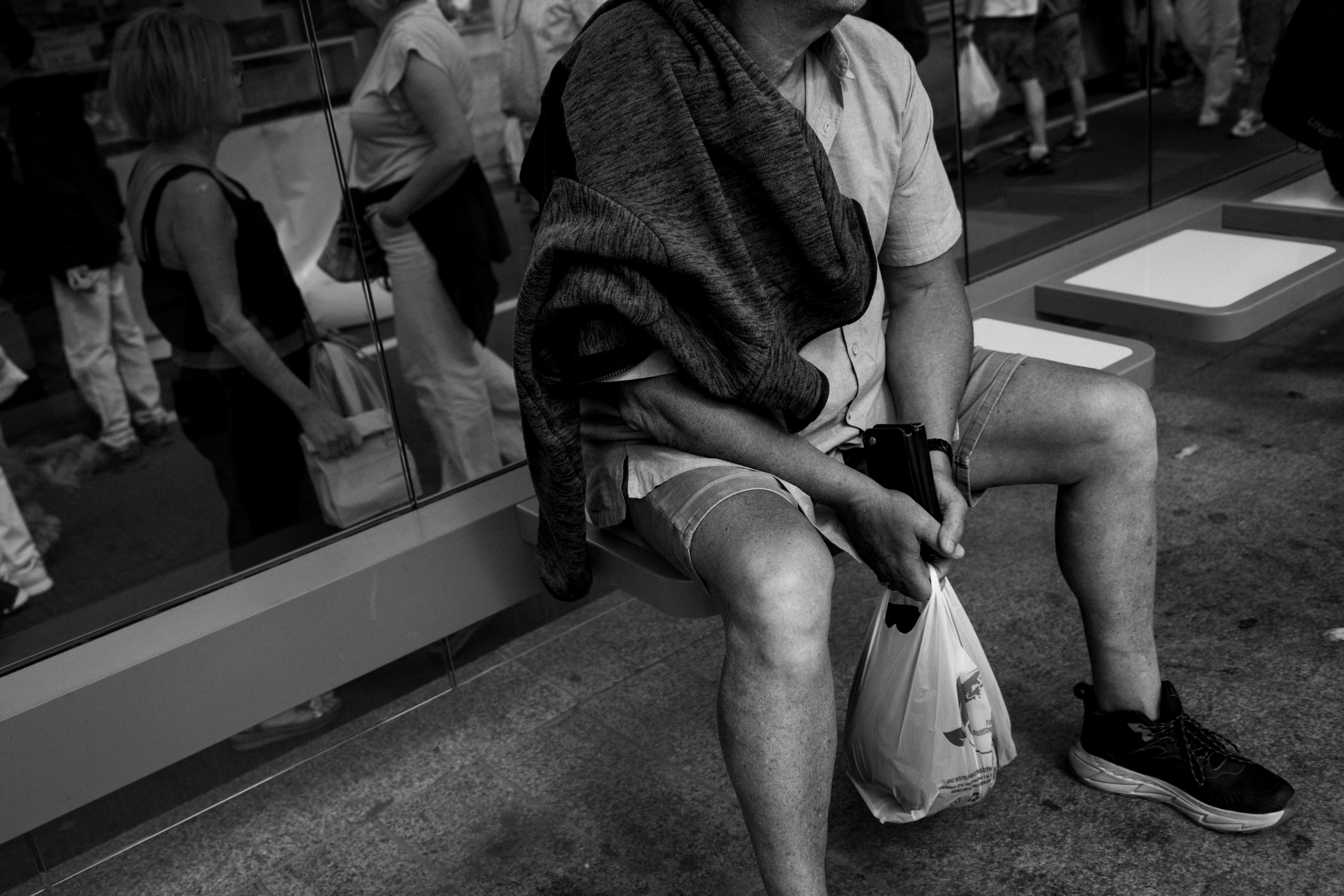 A person in shorts and a shirt sitting on a bench with a plastic bag