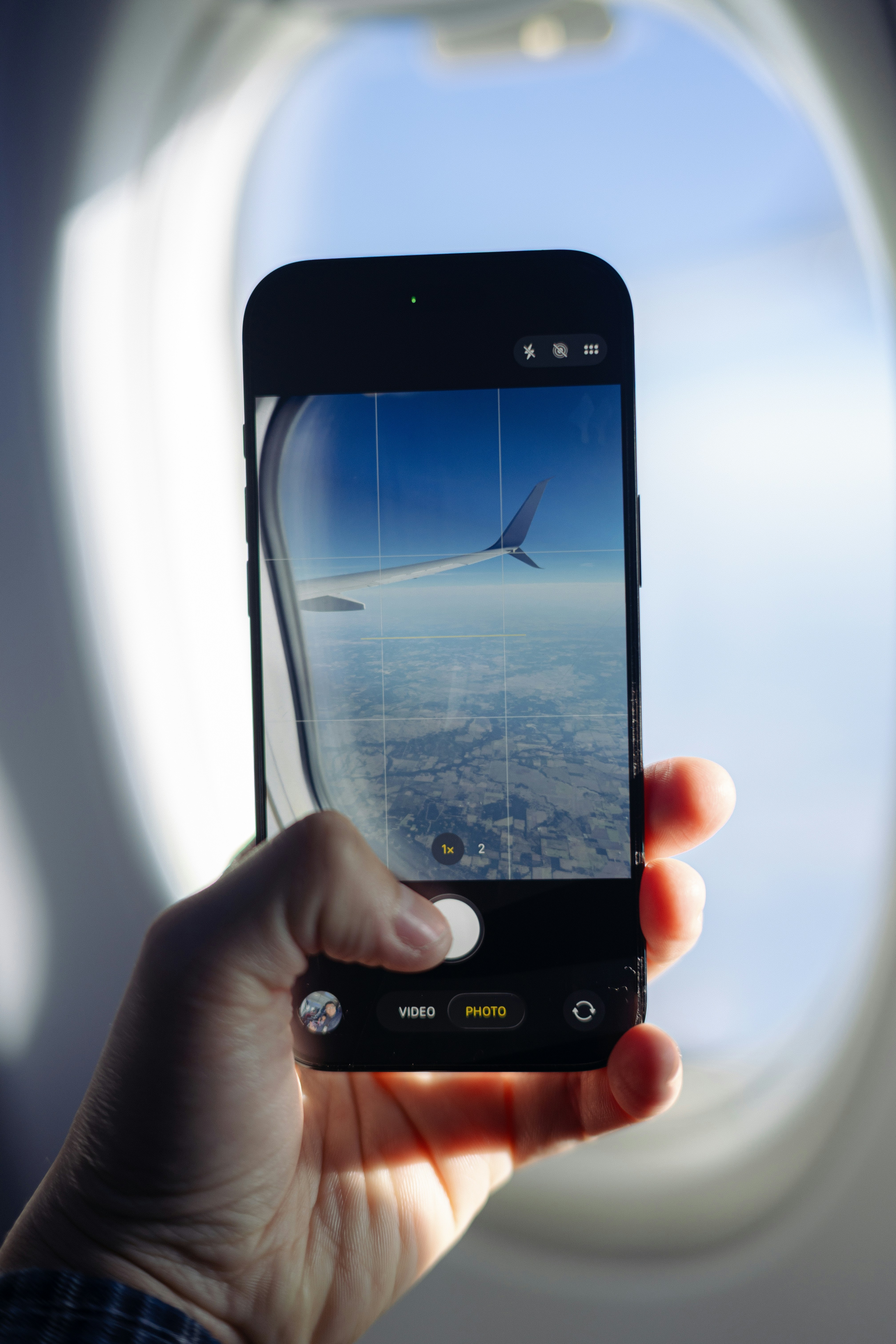 A hand holding a smartphone to photograph an airplane wing through a window