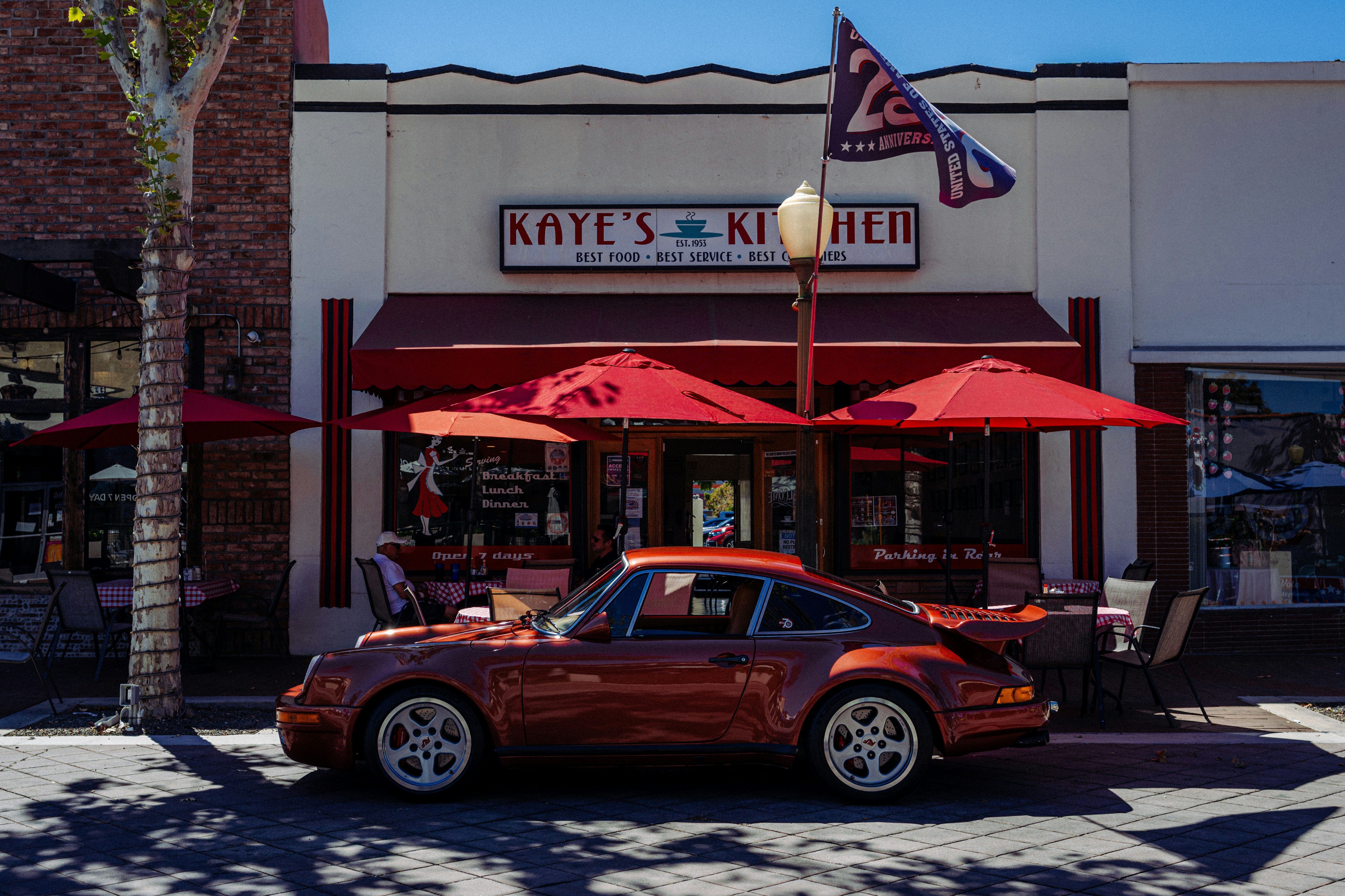 A vintage copper Porsche parked in front of Kaye's Kitchen with red umbrellas
