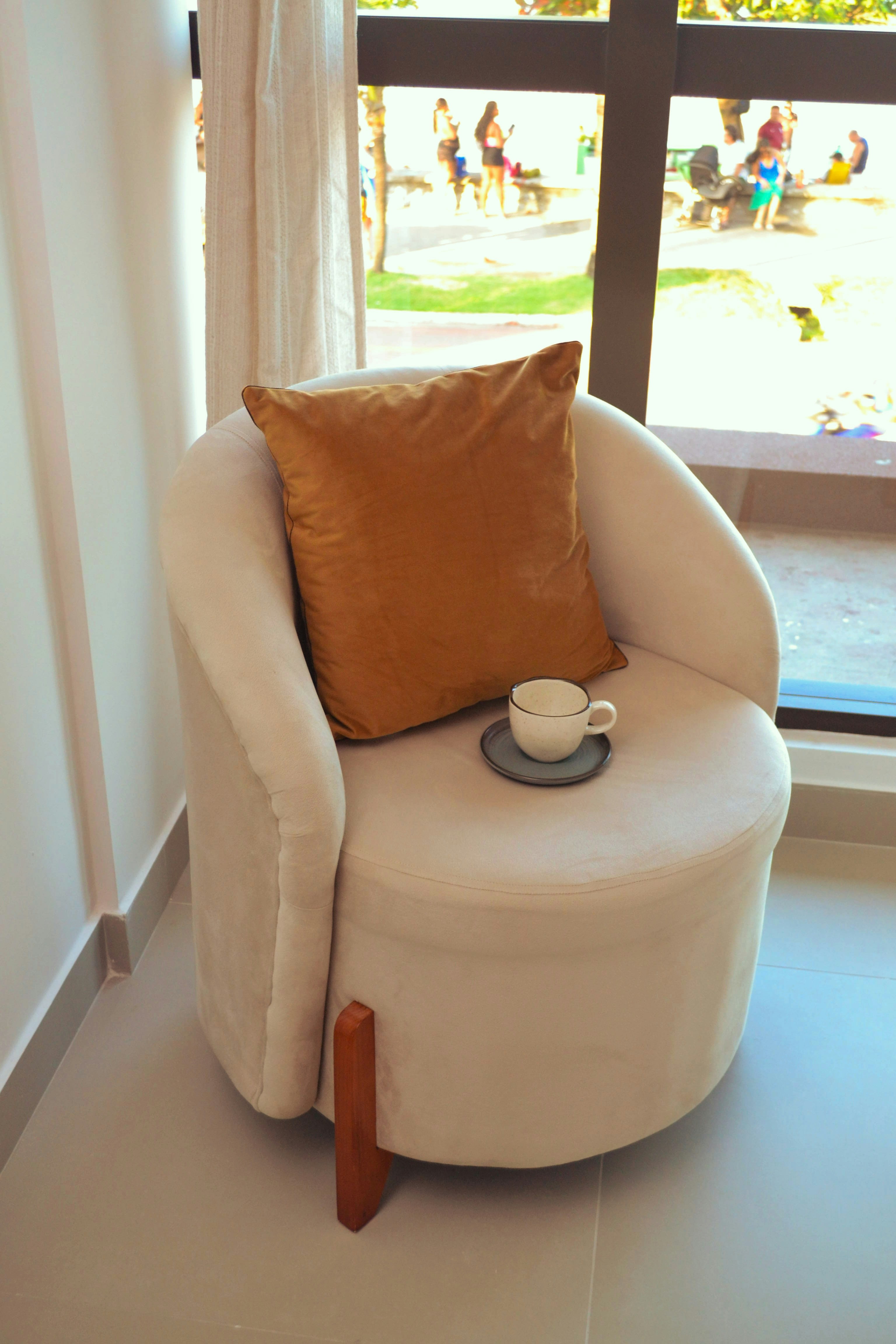 A cream armchair with a gold pillow and a small cup by a window