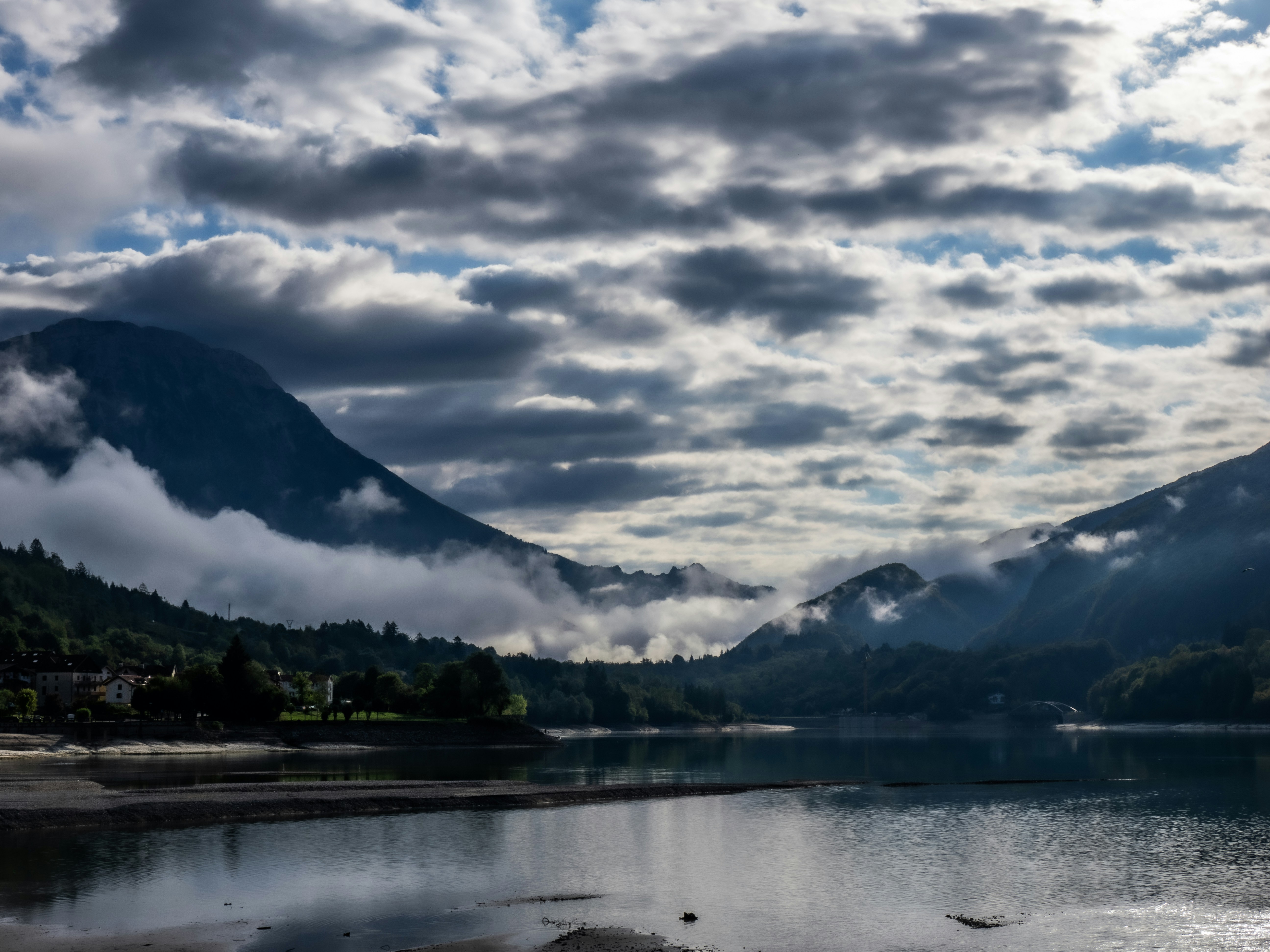 Low clouds drifting over a calm lake surrounded by mountains under a cloudy sky