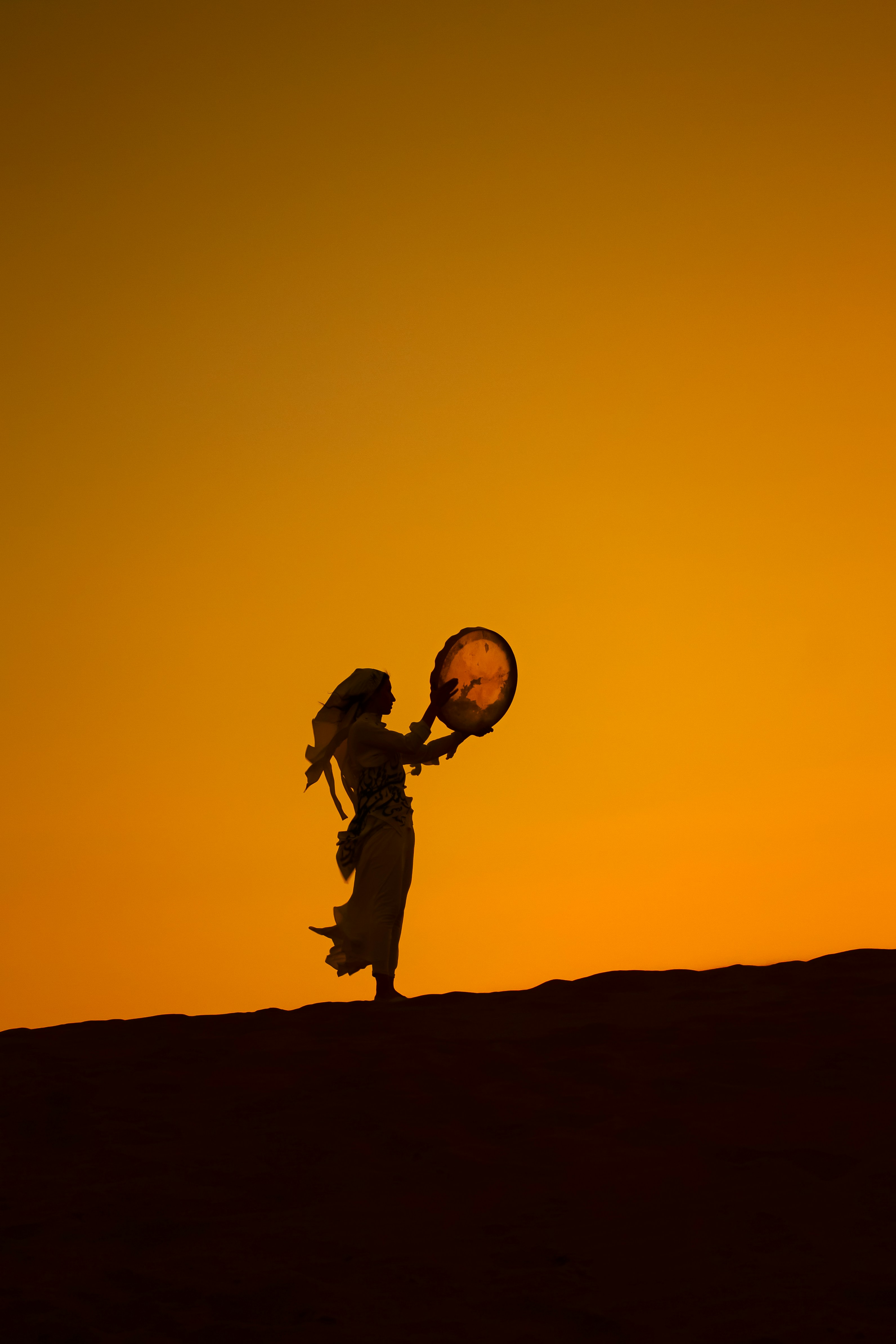 A person in silhouette holding a circular frame drum against a warm orange sky