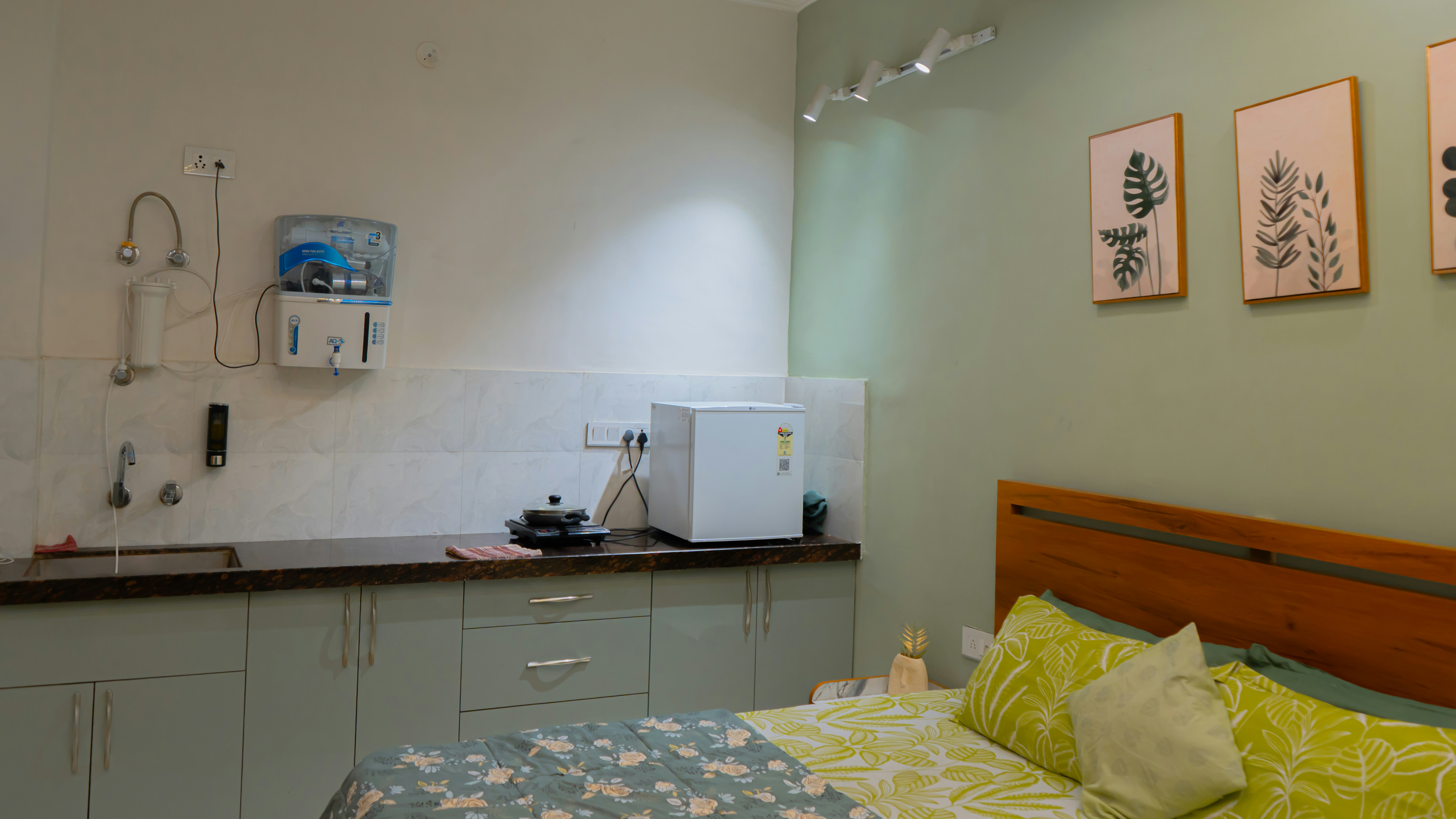Studio apartment with a double bed along one wall, a small kitchenette with a sink and mini-fridge beside it, and botanical prints on a green wall