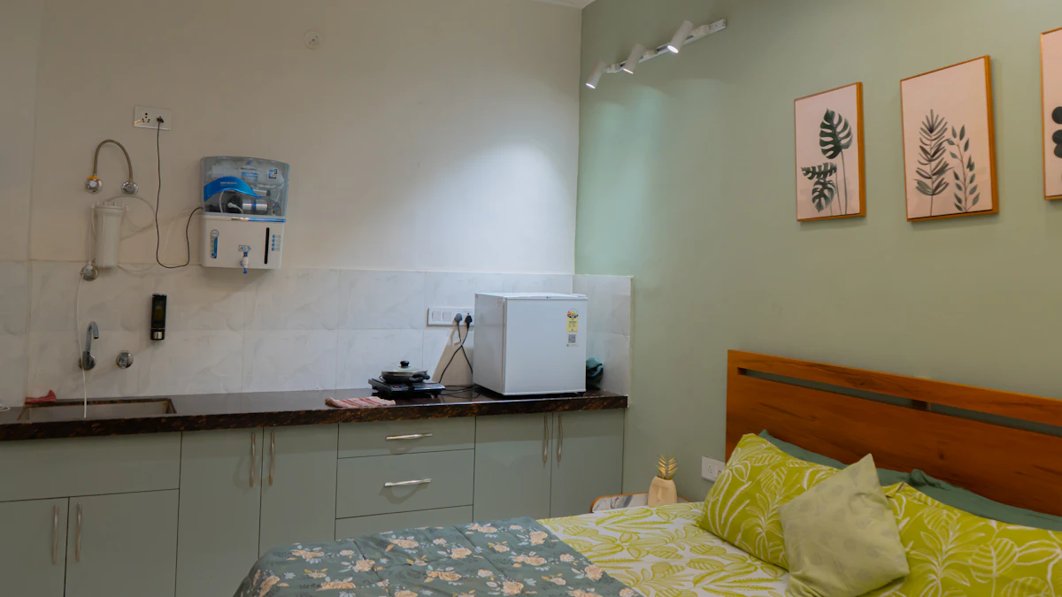 A small kitchenette with light green cabinets and a bed in a studio apartment