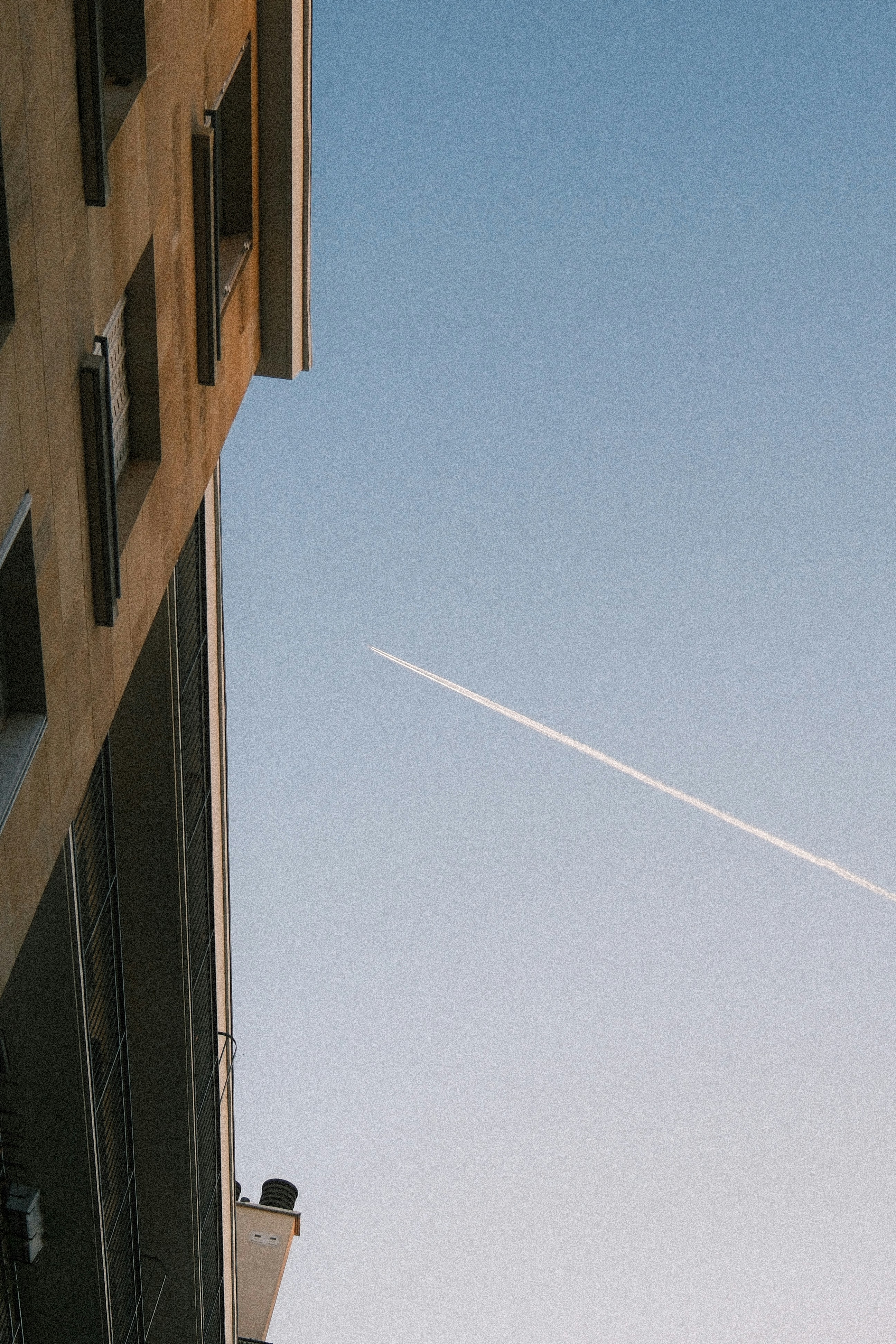 A vapor trail across a clear blue sky beside a tall stone building
