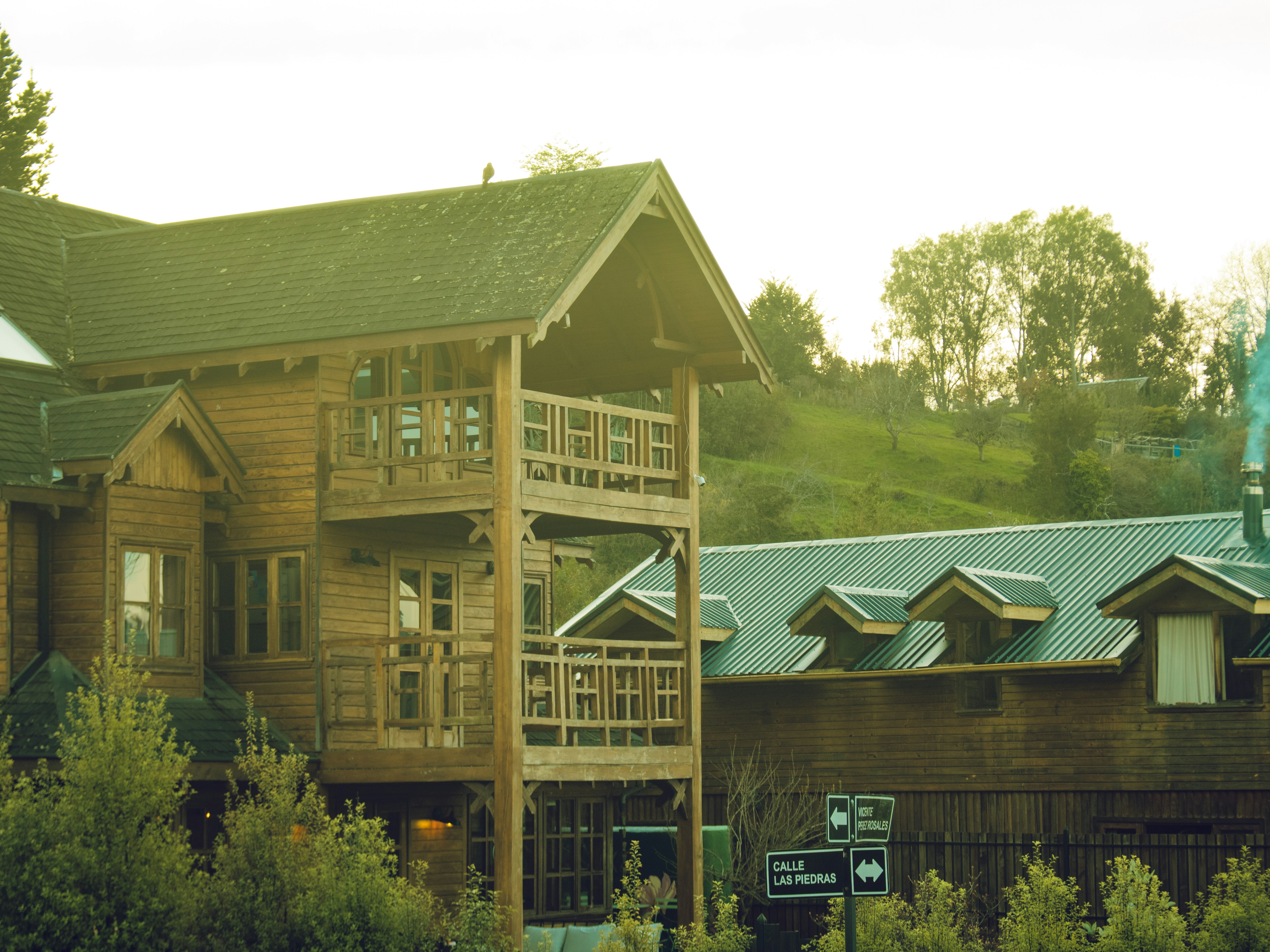 A wooden house with a balcony beside a building with a green metal roof