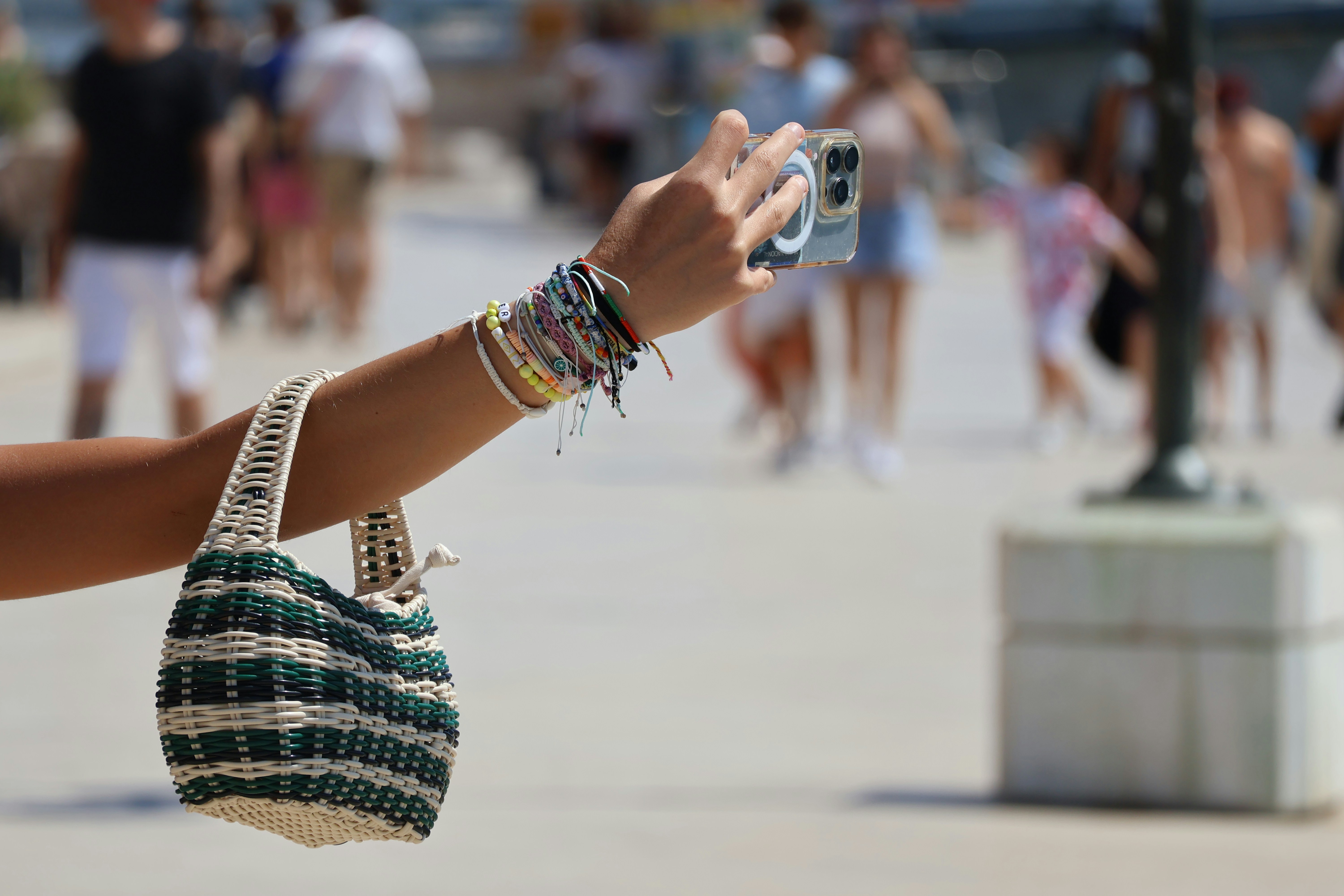 A person holding a smartphone and a woven bag on a sunny promenade