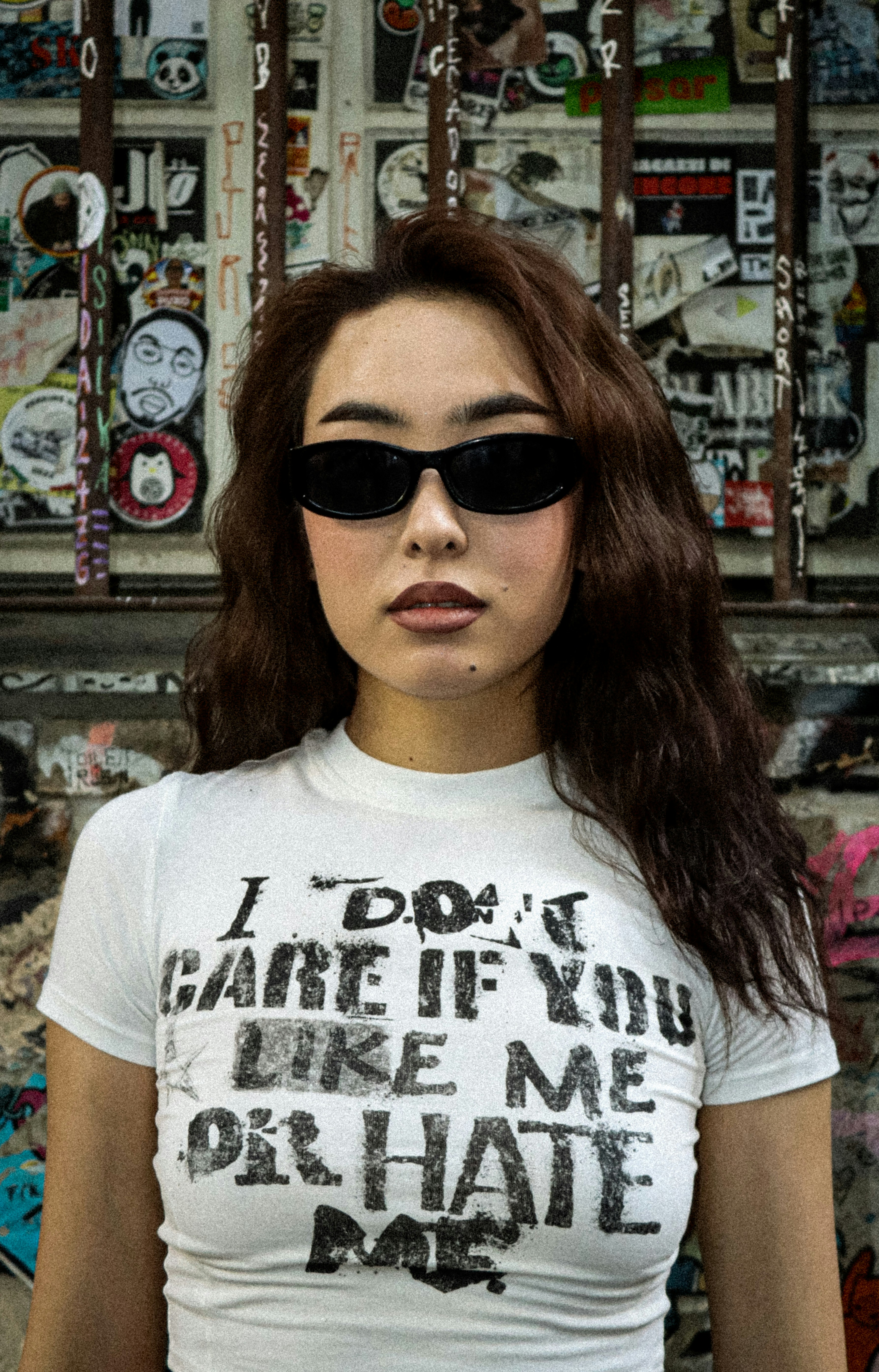 A woman wearing black sunglasses and a white t-shirt with text against a graffiti wall