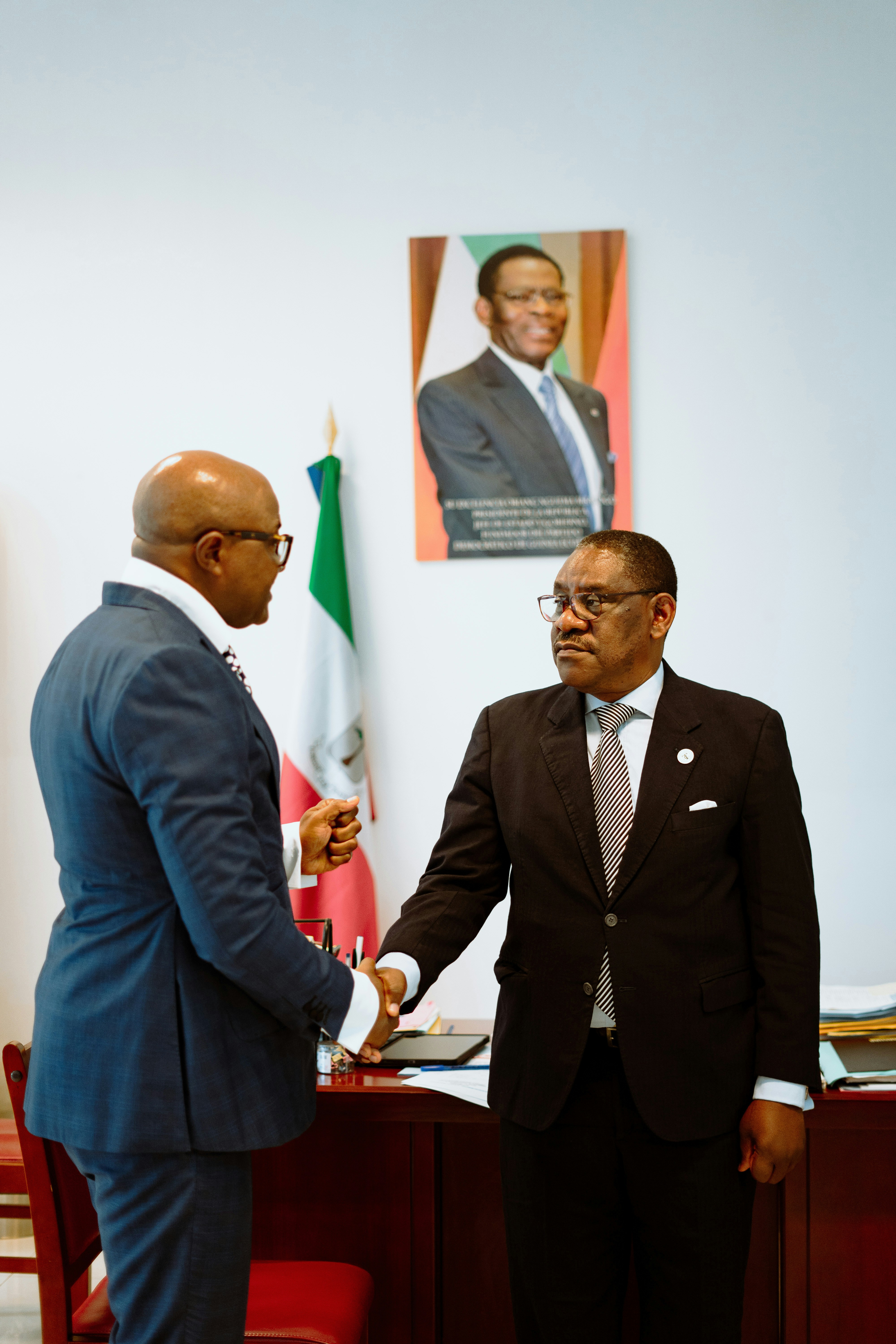 Two men in suits shaking hands in an office with a flag and portrait