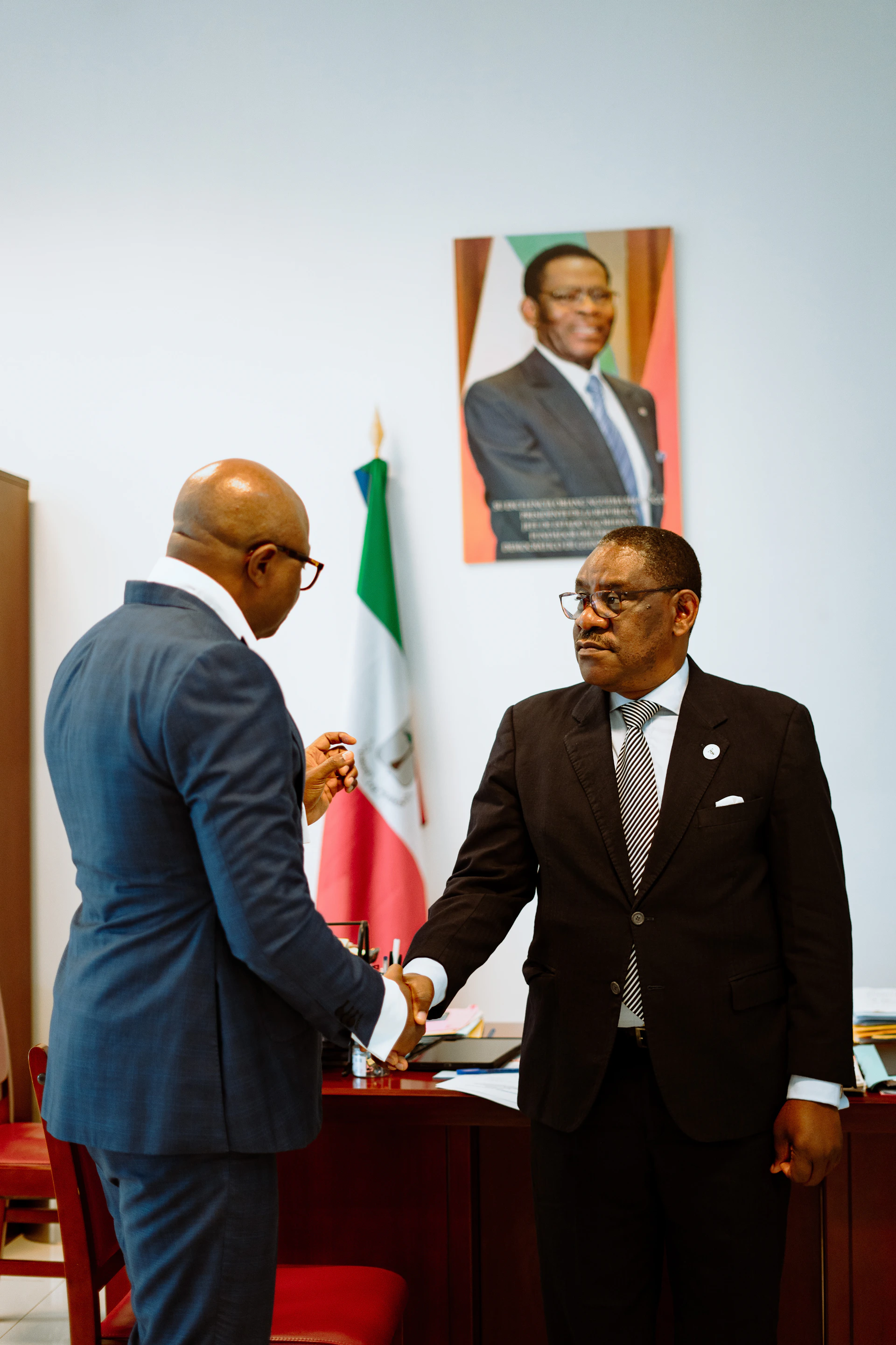 Two men in suits shaking hands in an office with a flag and portrait