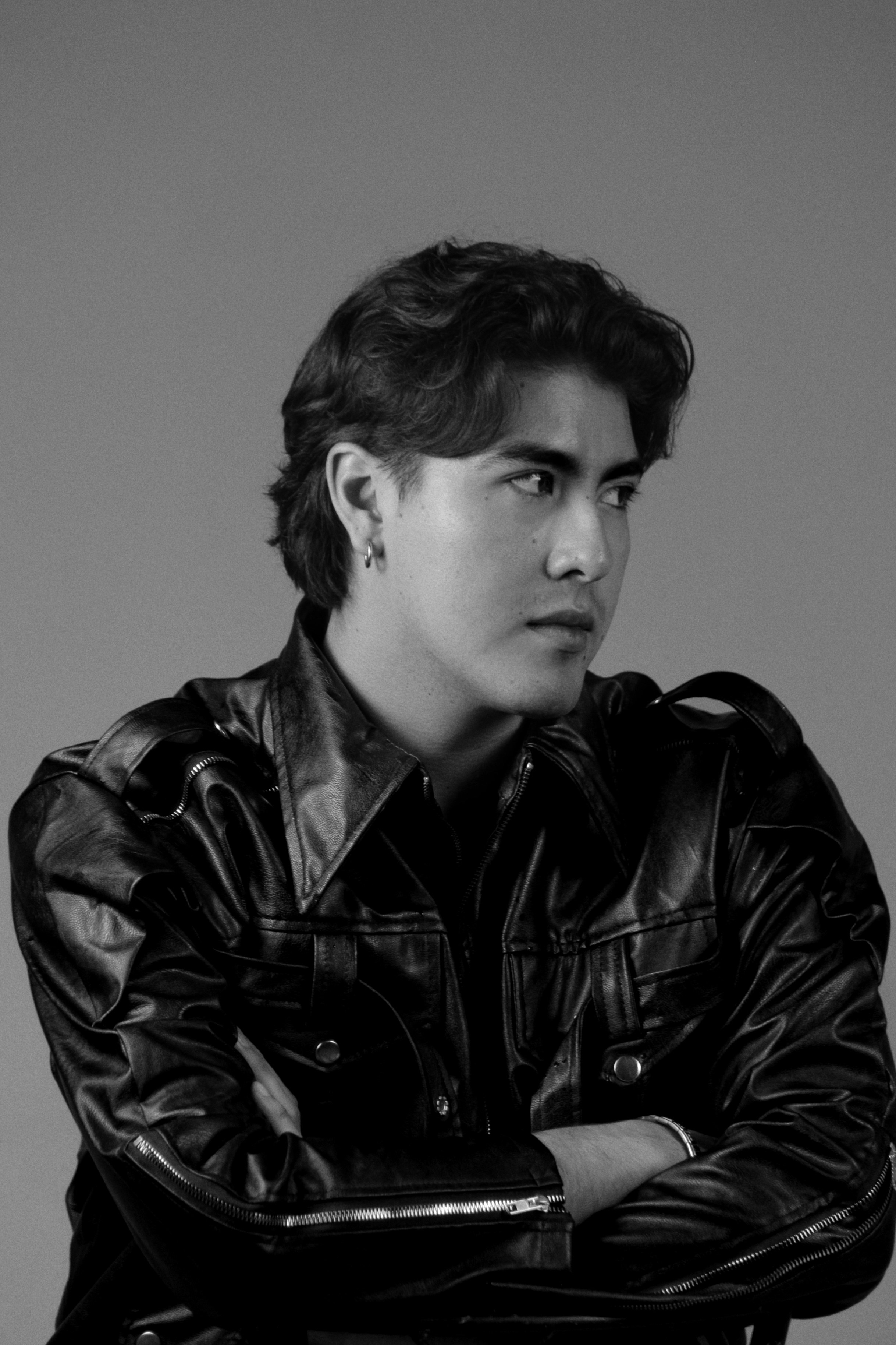 A man with wavy hair wearing a black leather jacket against a grey background
