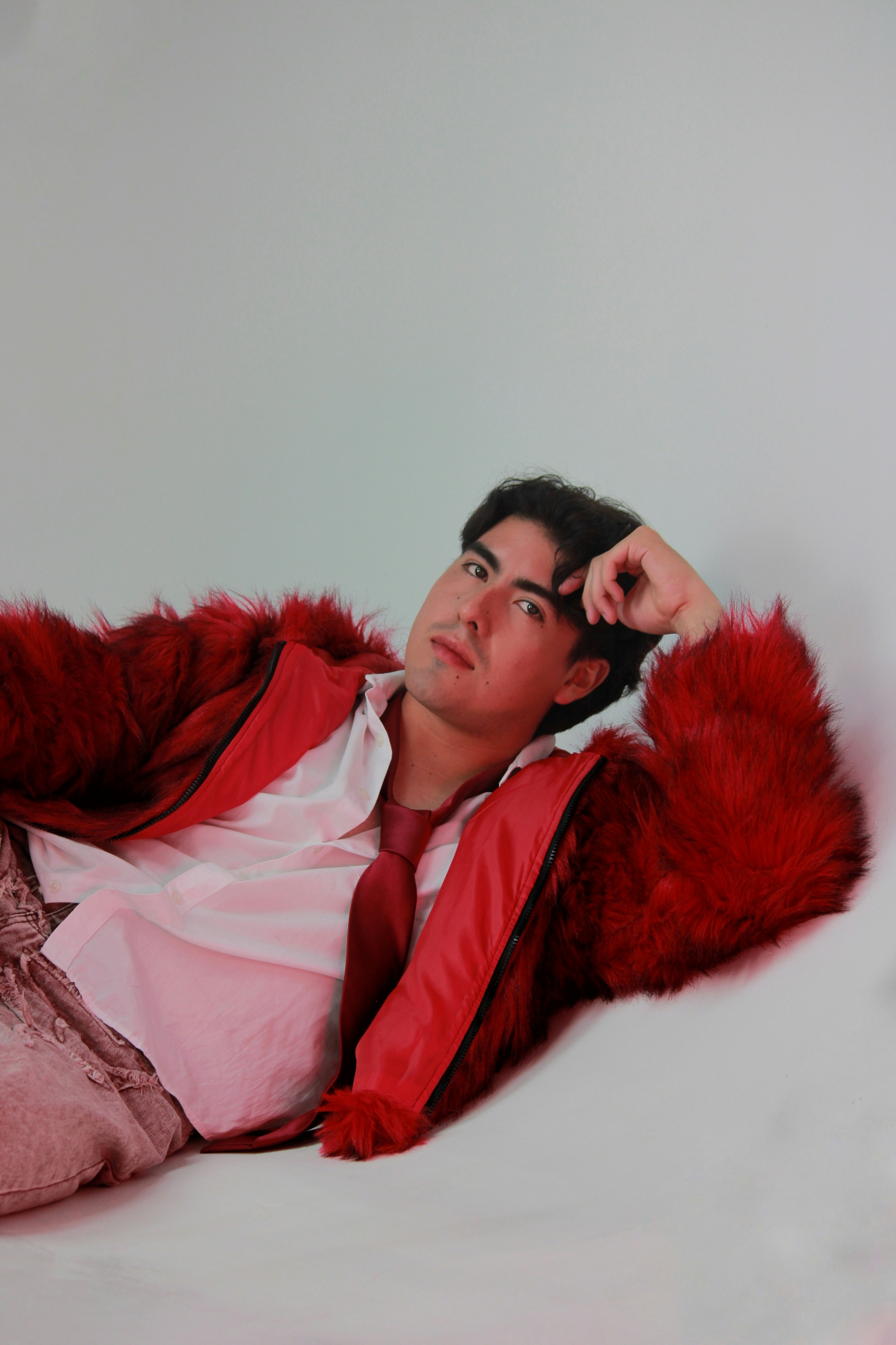 A man in a red faux fur jacket and tie reclining against a white background