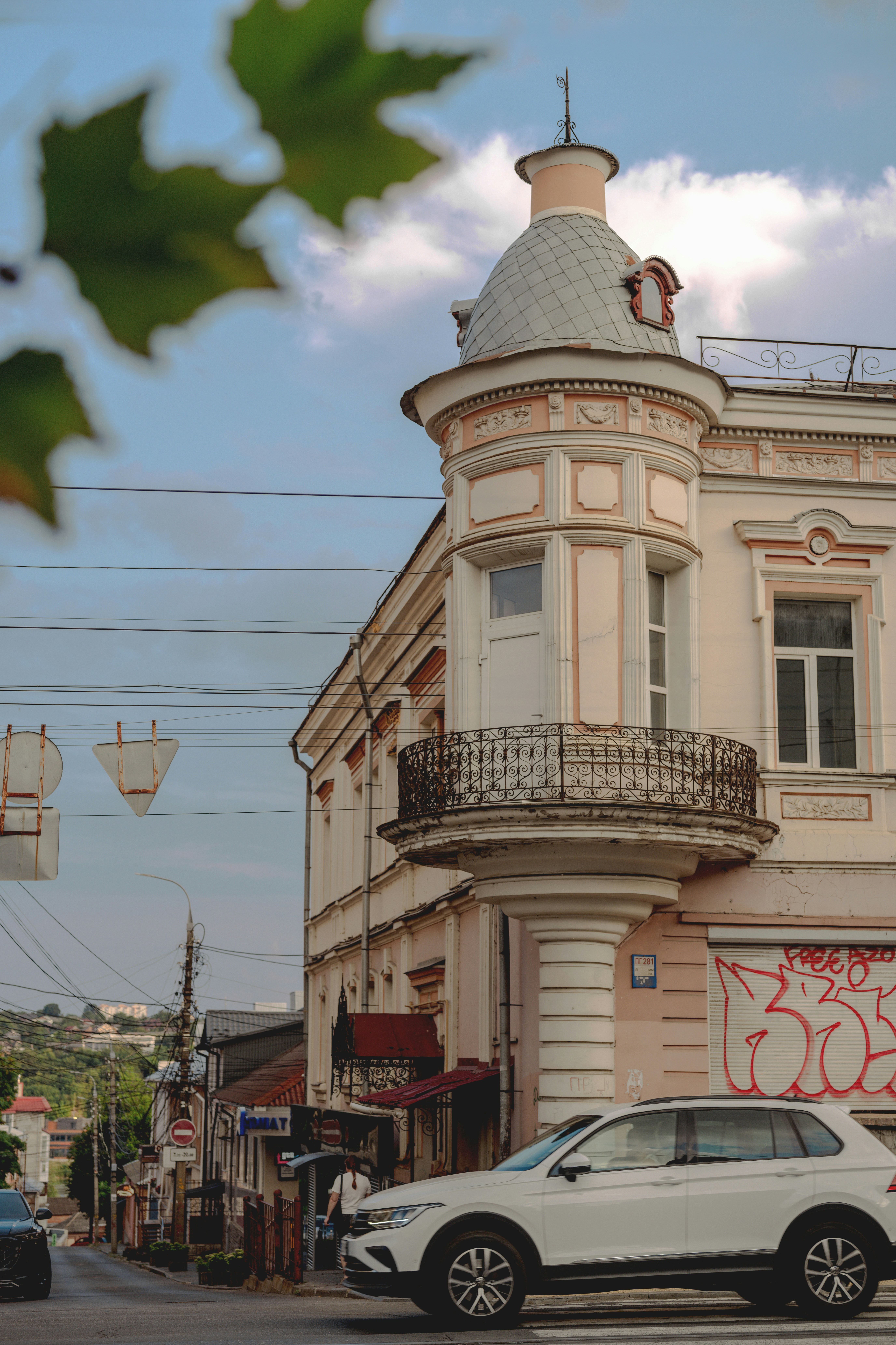 A white car driving past a historic building with a corner turret and balcony