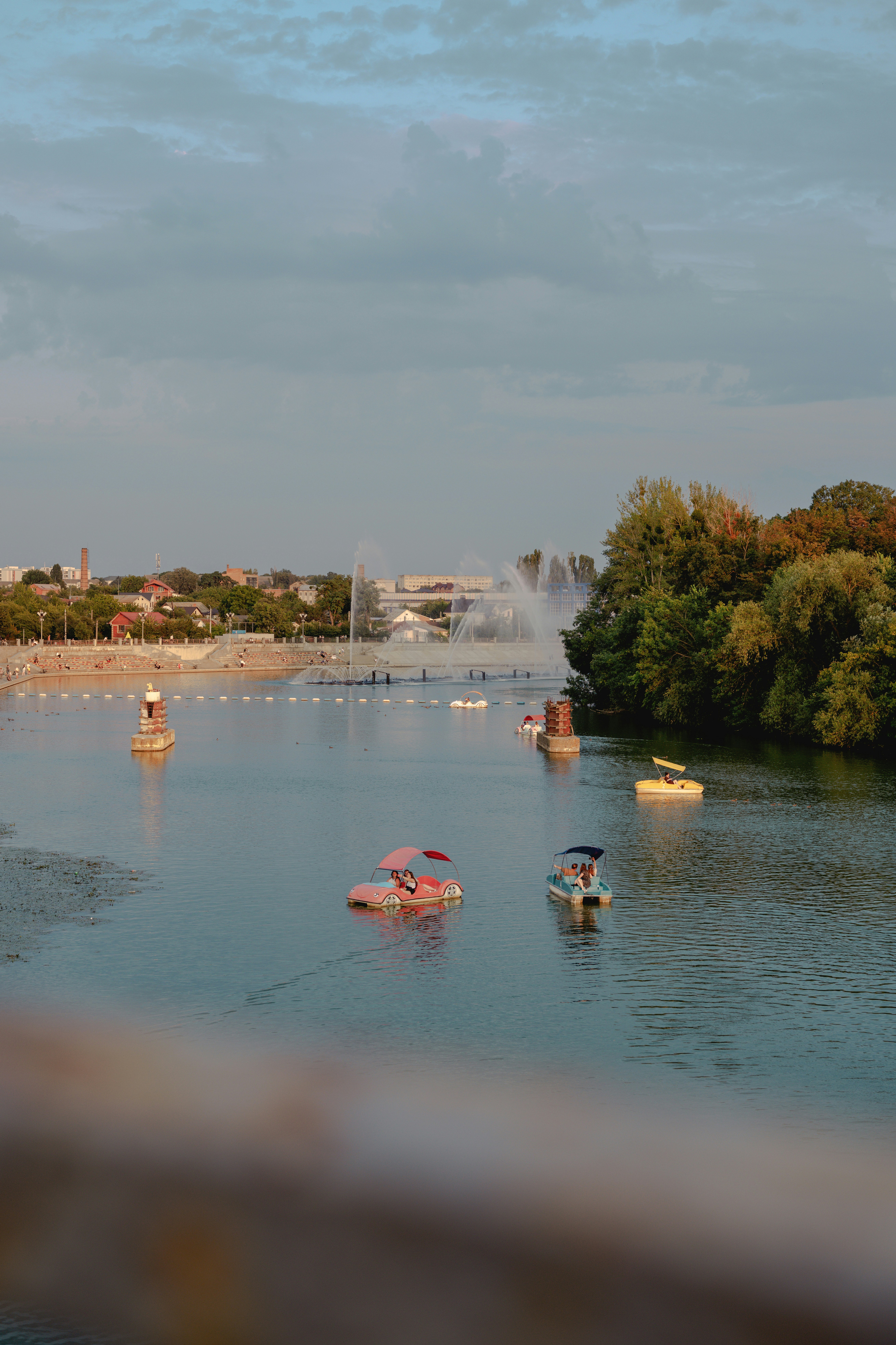 People in colorful pedal boats on a calm river with a distant fountain