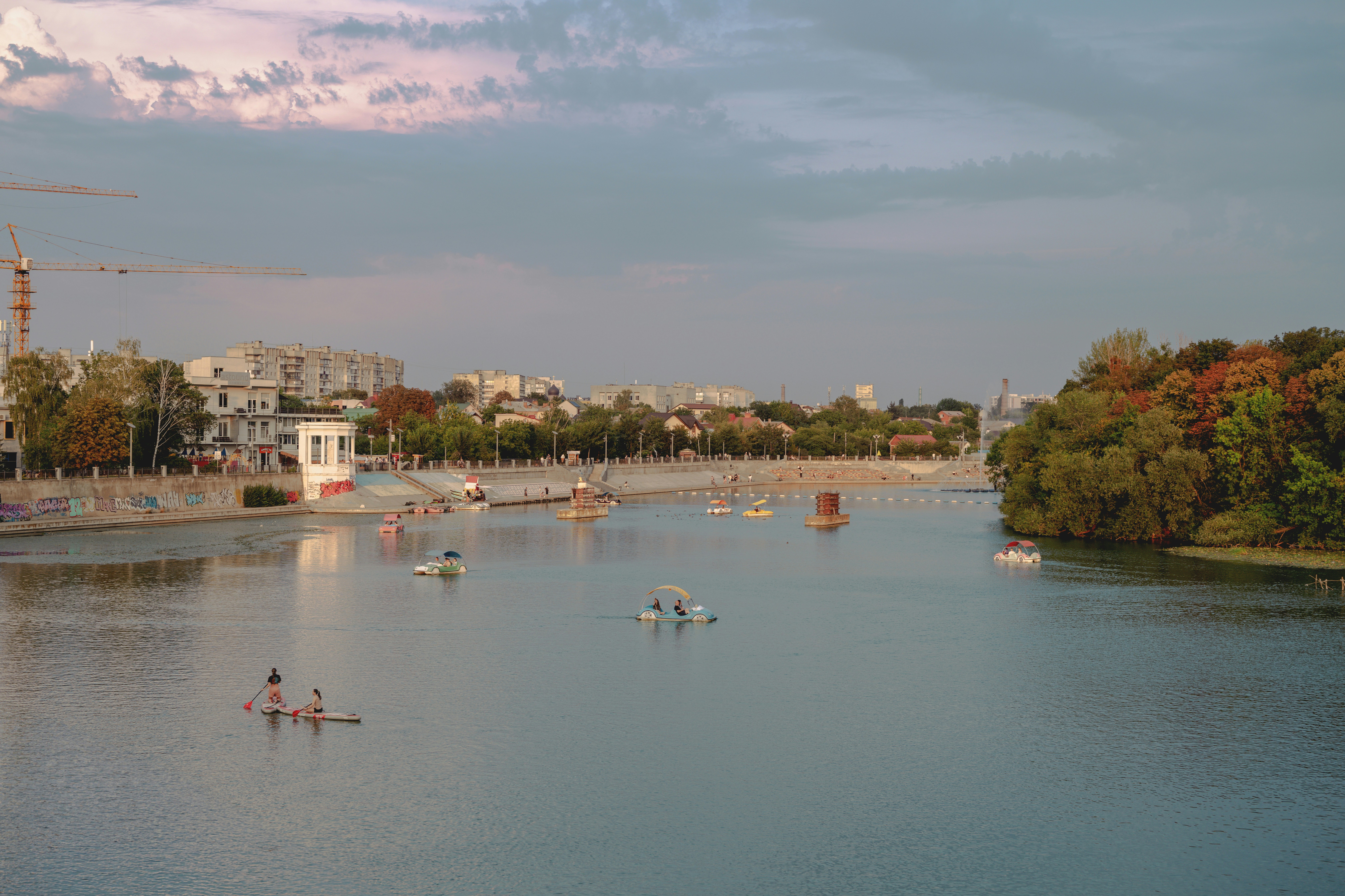 People on paddleboards and pedal boats on a river near a city embankment